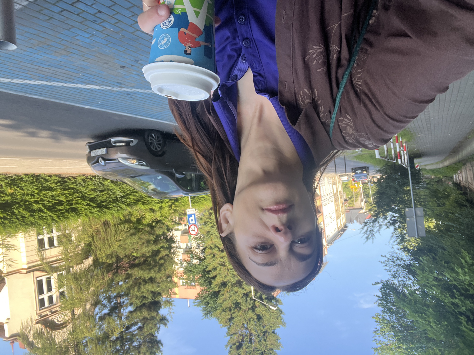 A woman with brown hair stands on a sidewalk, holding a coffee cup. She is wearing a brown patterned jacket over a purple collared shirt. The background shows a street with cars, buildings, and trees under a clear sky. A blue and white striped path is visible on the sidewalk in the foreground. The scene appears to be a city street during the daytime.