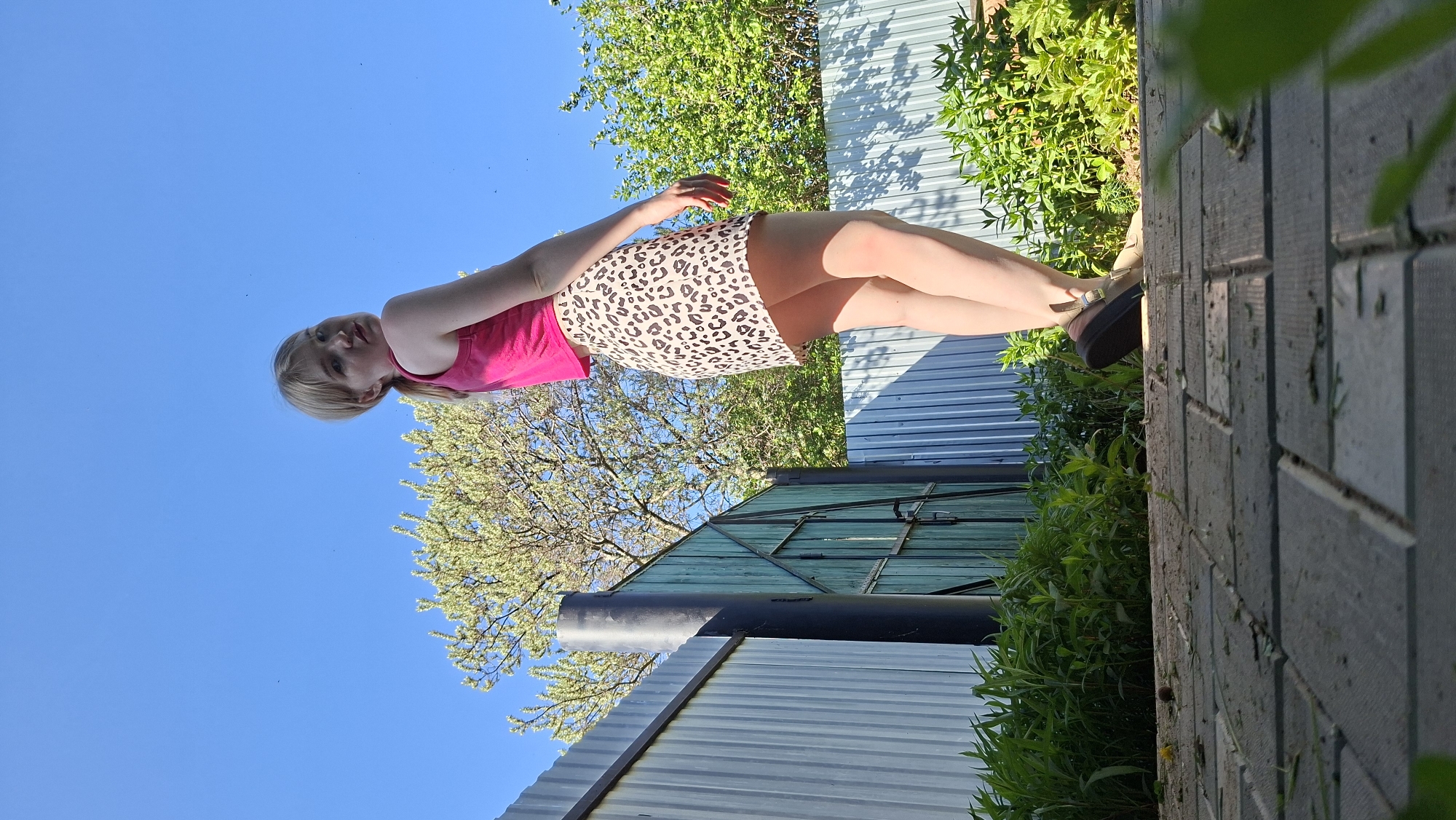 A photograph taken in Большой Букор, Russia, shows a young woman from a low angle. She is wearing a pink tank top and a short, leopard-print skirt. She also wears nude pantyhose and brown platform sandals. The sky is clear blue. Behind the woman, there is a corrugated metal fence and a green wooden shed with a black pipe. There are trees and foliage in the background. The ground is paved with gray bricks, and there are plants growing alongside it.