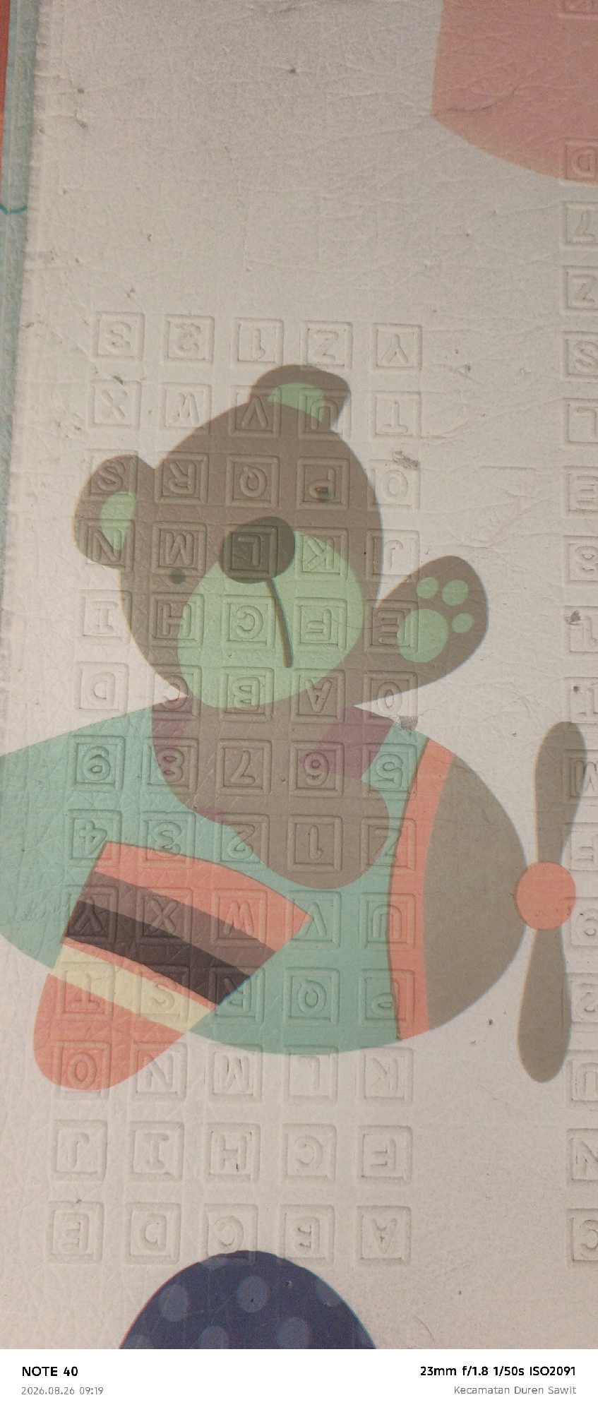 The image shows a close-up of a children's play mat with an embossed pattern of letters and numbers. A shadow of a cartoon teddy bear riding an airplane is cast over the mat. The play mat is textured and appears to be made of foam or rubber. The colors are muted, with the teddy bear and airplane shadow appearing in shades of brown and green. The background is a light, neutral color. In the bottom left corner, text reads "NOTE 40 2026.08.26 09:19". In the bottom right corner, text reads "23mm f/1.8 1/50s ISO2091 Kecamatan Duren Sawit". The location is specified as Kecamatan Duren Sawit, East Jakarta, Indonesia, suggesting this is a photograph taken within that administrative district. There are no people or animals visible in the image. The focus is on the texture and pattern of the play mat and the overlaying shadow.