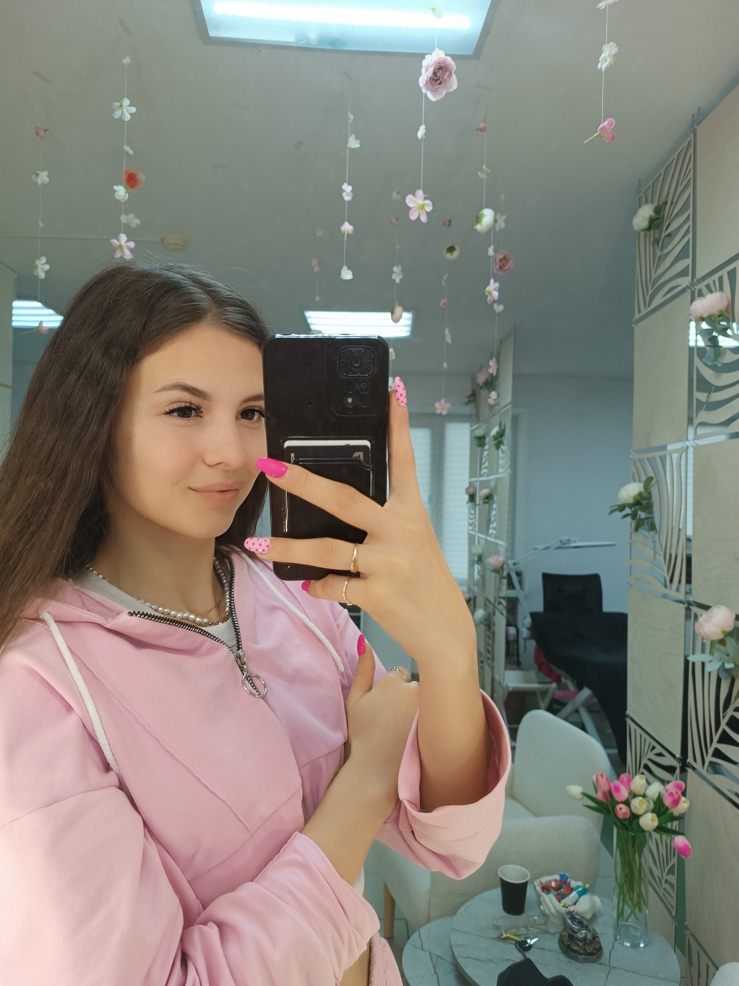A young woman is taking a selfie in a room decorated with floral elements. She is wearing a light pink hoodie over a white top, along with a pearl necklace. She has long brown hair and her nails are painted pink, with one nail featuring a pink polka dot design. She is holding a black smartphone. The room features hanging floral decorations and flowers arranged in vases. The location of this image is in the city of Unknown, Unknown.