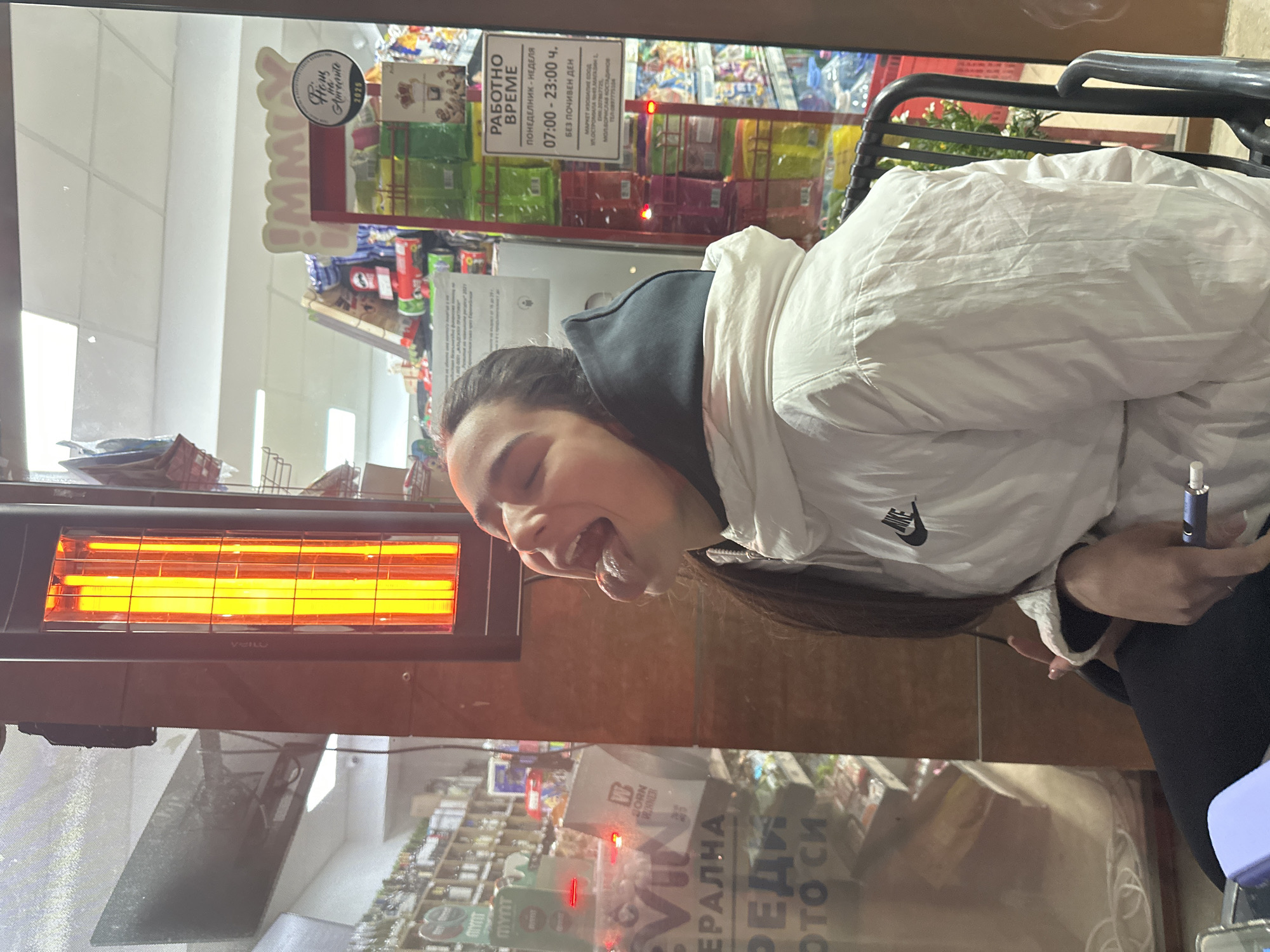 A young woman is sitting outside a shop, next to a VEILO brand electric heater with bright orange coils. She is wearing a white Nike jacket over a dark hoodie, has long dark hair, and is sticking her tongue out. She holds a blue electronic device in her right hand. The shop behind her has Bulgarian text on the window: "ЕРАЛНА РЕДИ ото си". A sign above the shop shelves reads "РАБОТНО ВРЕМЕ ПОНЕДЕЛНИК - НЕДЕЛЯ 07:00 - 23:00 ч. БЕЗ ПОЧИВЕН ДЕН" (Working Hours Monday - Sunday 07:00 - 23:00 without a day off). The sign also says "МАРКЕТ ИЗОБИЛИЕ БООД ... МАЛАДИН 1 МОЛ БОРИСЛАВ КОСТАДИНОВ". The location is Plovdiv, Bulgaria, specifically 1 Maladin Street, which corresponds to the address on the sign.