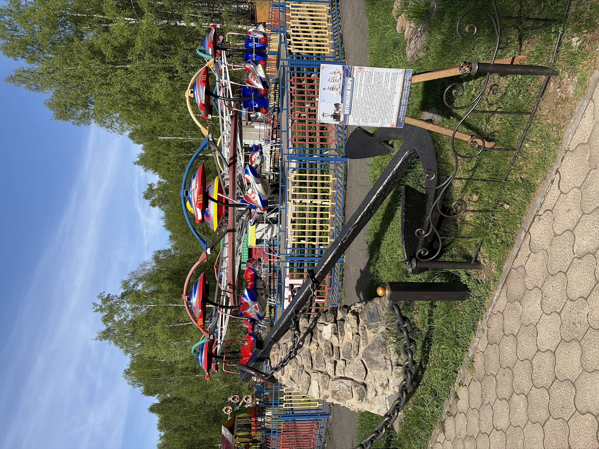 The image captures a view of the Votkinsk Anchor monument situated in Votkinsk, Russia. This monument features a large anchor, its chain leading to a stone base. Adjacent to the anchor is a sign in Russian, possibly detailing the history or significance of the anchor. Behind the monument, a children's ride with colorful, boat-like seats stands against a backdrop of trees and a blue sky. The foreground consists of a paved walkway and grassy areas.