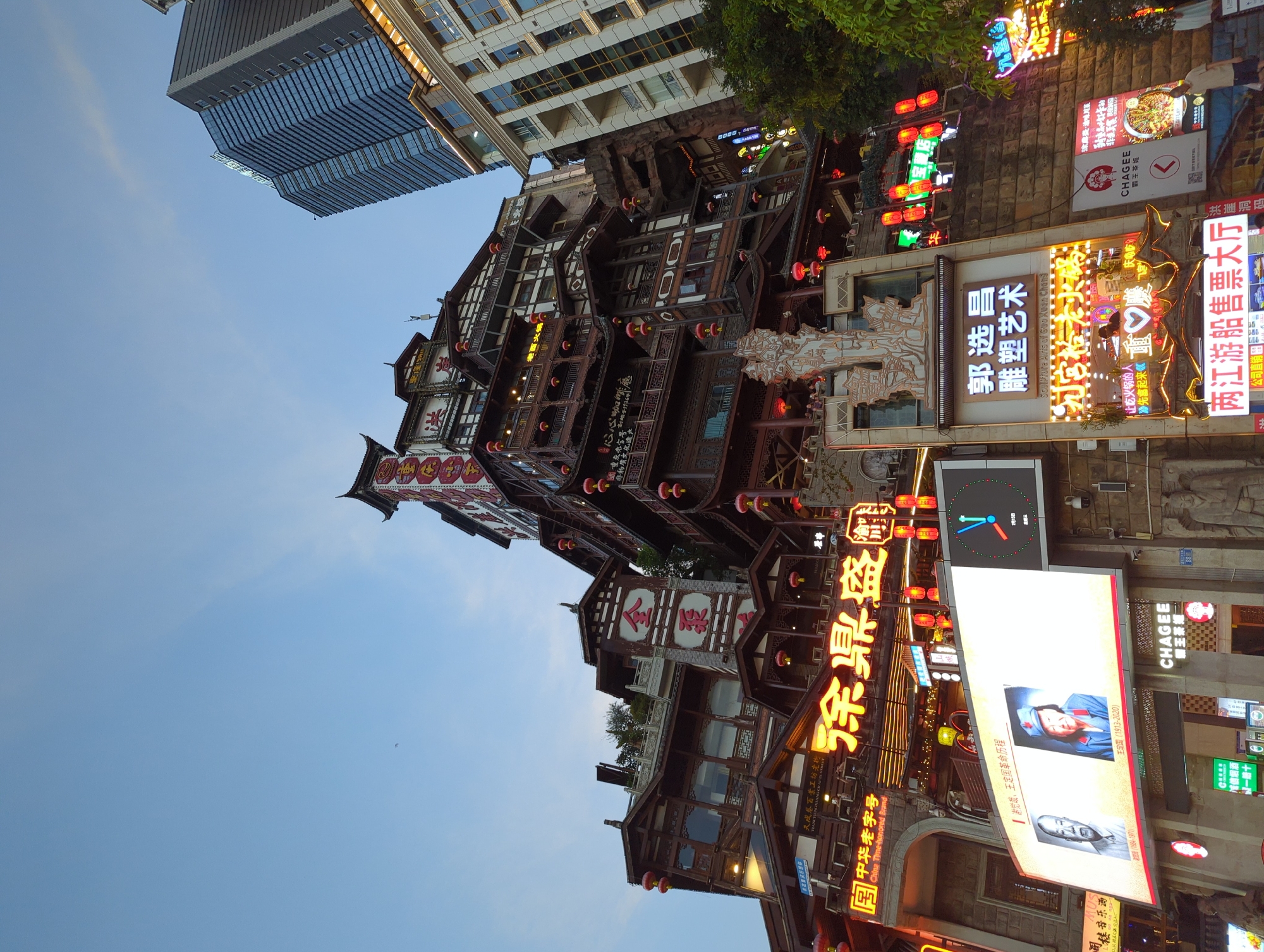 The image captures a street-level view of a bustling urban scene in Yuzhong District, China, likely in the evening given the illuminated signs. Dominating the frame is a multi-tiered, traditional Chinese architectural style building, reminiscent of a pagoda, adorned with numerous red lanterns and ornate carvings. This structure is juxtaposed with a modern, glass-fronted skyscraper in the background, creating a striking contrast between old and new.
In the foreground, a large, brightly lit digital billboard displays two portraits. The left portrait shows an elderly man, identified as "Mao Zedong (1893-1976)". The right portrait depicts an elderly woman in a red hat and uniform, identified as "Jiang Qing (1913-1991)". Below these portraits, text indicates "Chairman, Founding of the Nation Revolutionary Period." Another digital screen displays a clock with Chinese characters, possibly indicating time or a countdown.
Adjacent to the billboard are various neon signs in Chinese, advertising shops and services. One sign in yellow characters reads "徐鼎盛" (Xu Ding Sheng), which could be a business name. Another sign, in white characters against a dark background, proclaims "郭选昌 雕塑艺术" (Guo Xuan Chang Sculpture Art), indicating a sculpture exhibition. Below this, more illuminated signs feature neon lights in various colors, including hearts and slogans.
Further down the street, other signs are visible, including one for "CHAGEE 霸王茶姬" (Ba Wang Cha Ji), a tea shop. A person, likely a male, is seen walking in the lower right corner of the image. The street is paved, and the buildings are constructed from stone and wood. The sky above is a clear blue, suggesting it is either dusk or dawn. The overall impression is one of a vibrant, commercial area blending historical and contemporary elements.