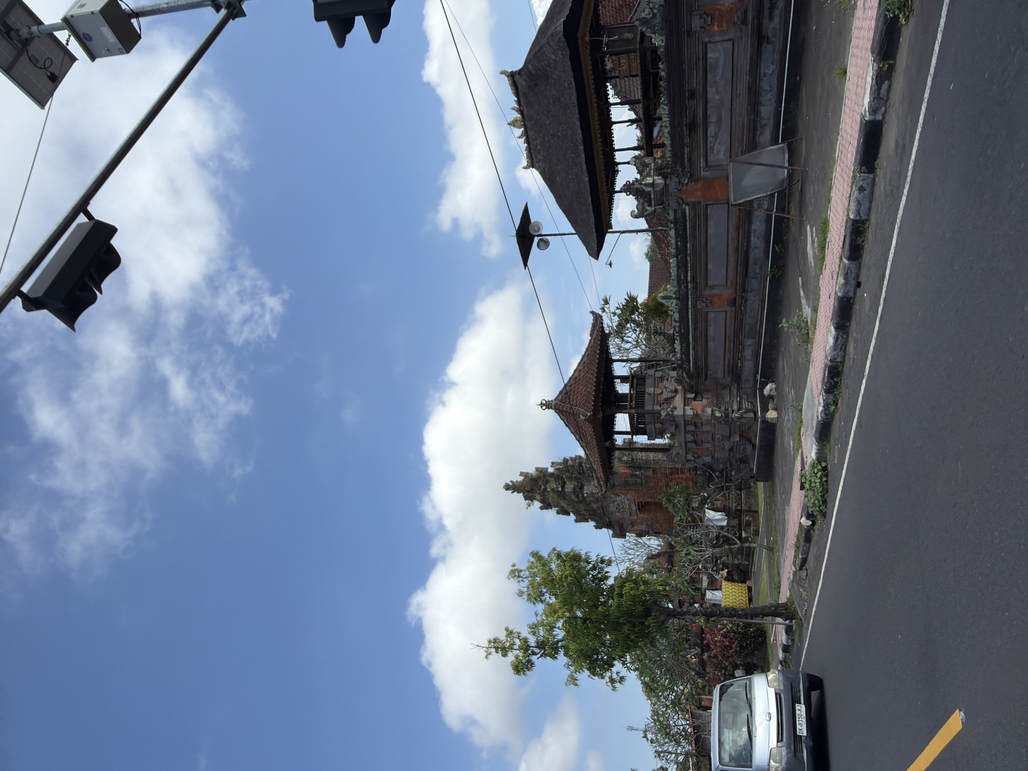 In Blahbatuh, Indonesia, a silver minivan with license plate DK 8150 KJ drives on a road past a traditional Balinese building complex. The building features intricate stone carvings and multiple thatched-roof structures, one with an ornate apex. Adjacent to the buildings is a sidewalk with decorative patterned tiles and large stone borders. A solitary green tree stands to the left of the building complex. Overhead, a clear blue sky is dotted with white clouds. Traffic lights and solar panels are visible on poles along the road.