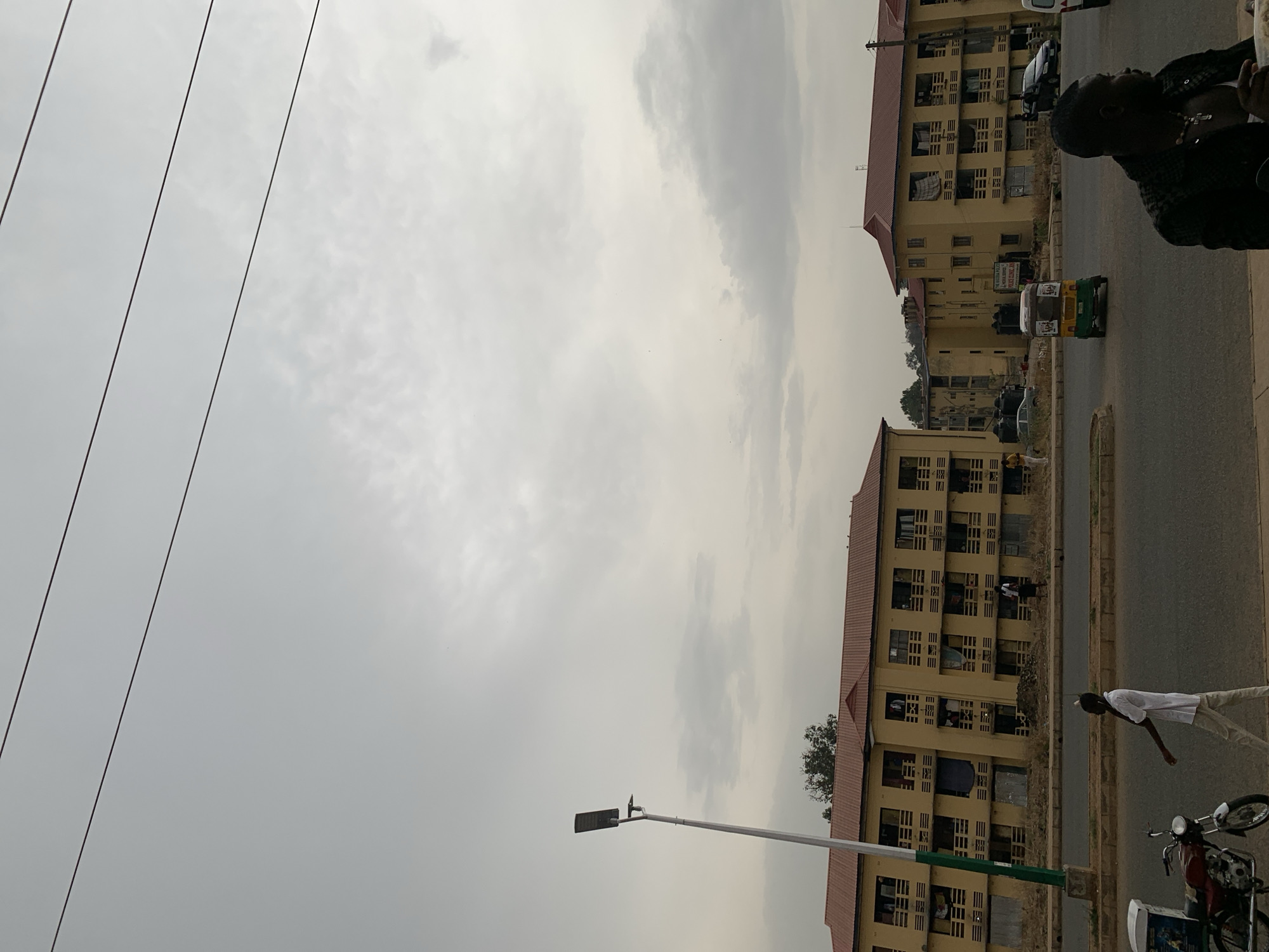 The image depicts a street scene in Jos, Nigeria. There are two large, multi-story buildings with yellowish facades and red roofs on either side of the road. The buildings appear to be residential or institutional. The sky is overcast. A three-wheeled vehicle (keke) and several cars are on the road. At least two people are visible, one on the right side of the image and another closer. A street lamp stands near the bottom left corner of the image. Power lines run across the upper left portion of the frame. A parked motorcycle and other objects are visible on the right side of the street.