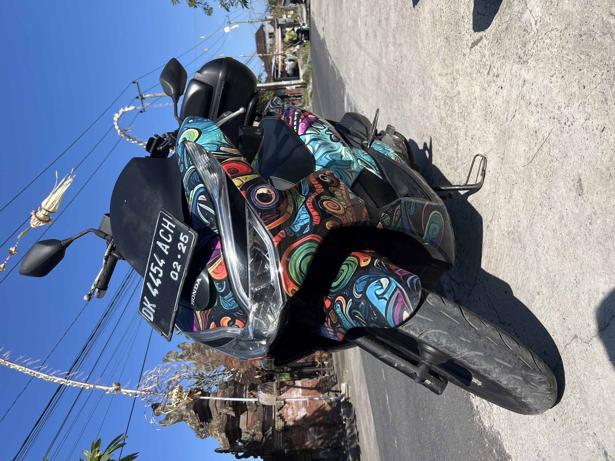 A black Honda scooter with a vibrant, colorful graphic wrap is parked on a concrete surface in Sukawati, Indonesia. The scooter's license plate reads "DK 4454 ACH 02-25". In the background, a traditional Balinese stone temple structure is visible to the left, with hanging decorative elements and power lines overhead. A street stretches into the distance, with houses and a few people on motorcycles. The sky is clear and blue.