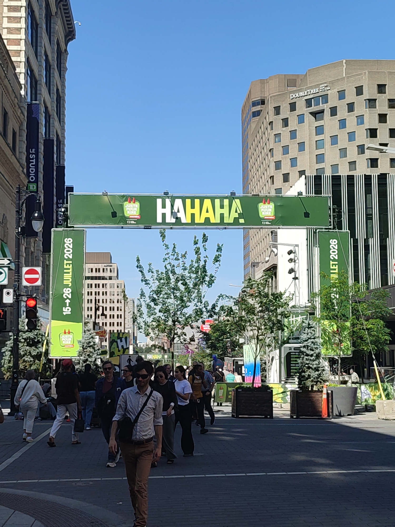 In Montreal, Canada, a street scene unfolds under a clear blue sky, dominated by a large green banner that reads "HAHAHA" in bold yellow letters, suggesting a festive or comedic event. The banner is suspended over a street, likely a commercial district, indicated by the surrounding buildings. To the left, a tall, multi-story building with a brick facade and numerous windows is visible. To the right, a modern, grey building with a similar grid-like window pattern rises above.
The street is paved with cobblestones, and several people are walking in various directions. In the foreground, a man wearing sunglasses and a light-colored collared shirt with dark pants walks towards the viewer, carrying a dark messenger bag over his shoulder. Behind him, other pedestrians are seen in groups and individually, some appearing to be socializing or looking at their phones.
On the left side of the street, traffic signals are present, with a red light illuminated. A circular red and white "stop" sign is also visible. Vertical green banners with French text, including "15-26 JUILLET 2026," and "JUSTE POUR RIRE," are attached to poles flanking the main banner, identifying the event as "Just for Laughs" comedy festival, occurring from July 15-26, 2026. Small evergreen trees in decorative planters are strategically placed along the sidewalk, adding a touch of greenery to the urban environment. The overall impression is of a vibrant street, likely a pedestrian area or a street closed for an event, with a clear emphasis on the upcoming comedy festival.