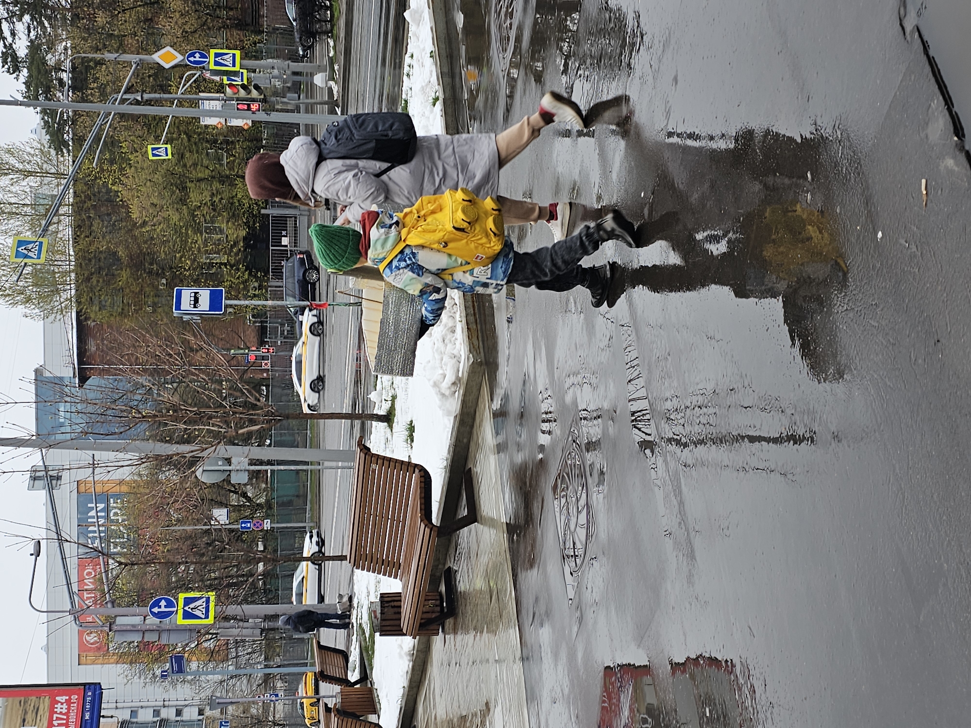 An adult and a child are depicted walking on a wet urban sidewalk, seen from behind. The image is oriented vertically (rotated 90 degrees clockwise). The adult wears a light grey coat, a dark backpack, and light-colored pants, with their head covered by a brown hood or hat. The child, slightly ahead, is dressed in a patterned blue and white jacket, a bright yellow backpack designed with prominent stud-like elements (reminiscent of LEGO), dark pants, black boots, and a green knit hat.
The ground is covered in puddles and reflective wet pavement, clearly showing the reflections of the figures. Patches of snow are visible on the narrow grassy strips alongside the sidewalk, indicating cold or cool weather conditions with recent rain or snowmelt. Several brown wooden benches are placed along the sidewalk.
In the background, an urban street with passing cars is visible, along with buildings, some with large windows. Numerous street signs are mounted on poles, including multiple pedestrian crossing signs (both blue square and yellow diamond variants), a blue bus stop sign, a yellow yield sign, and a blue "no parking" sign. Traffic lights are also present, one displaying a red signal.
Visible text includes "17#4" and "МS 19777 B" on a red sign at the bottom left, and faint Cyrillic lettering, including "LEINN," on a banner in the background. The scene suggests a typical urban setting in Khimki, Russia.