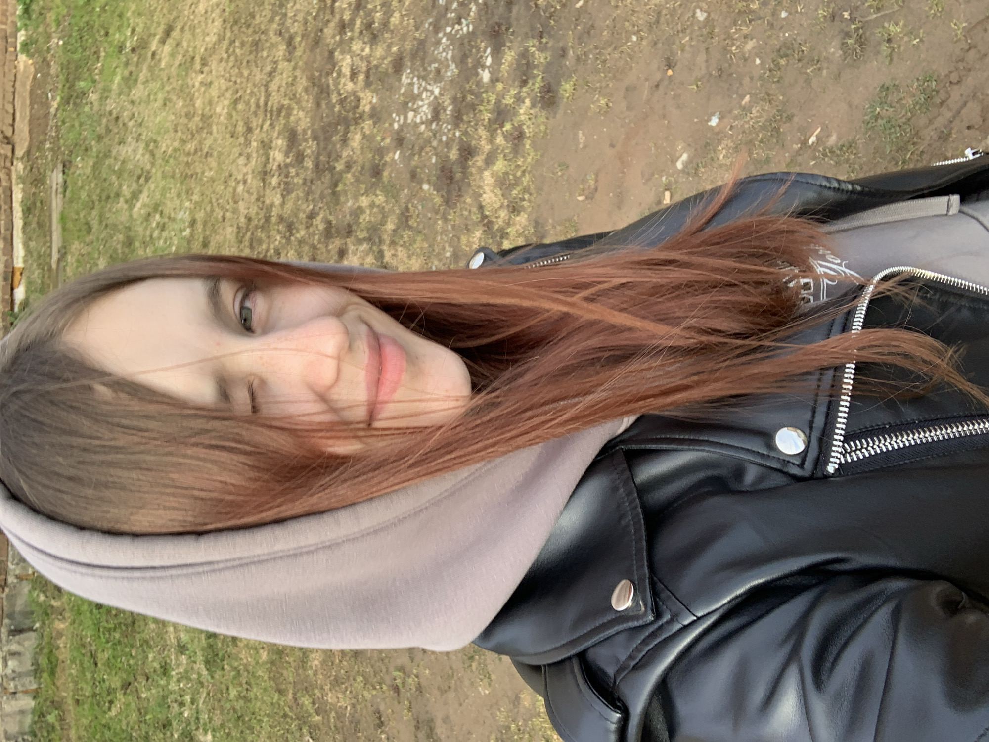 A young woman with brown hair and light complexion is facing the camera with a slight smile. She is wearing a grey hoodie and a black leather jacket. The background is a patch of grass and dirt. The location is Фоки, Russia.