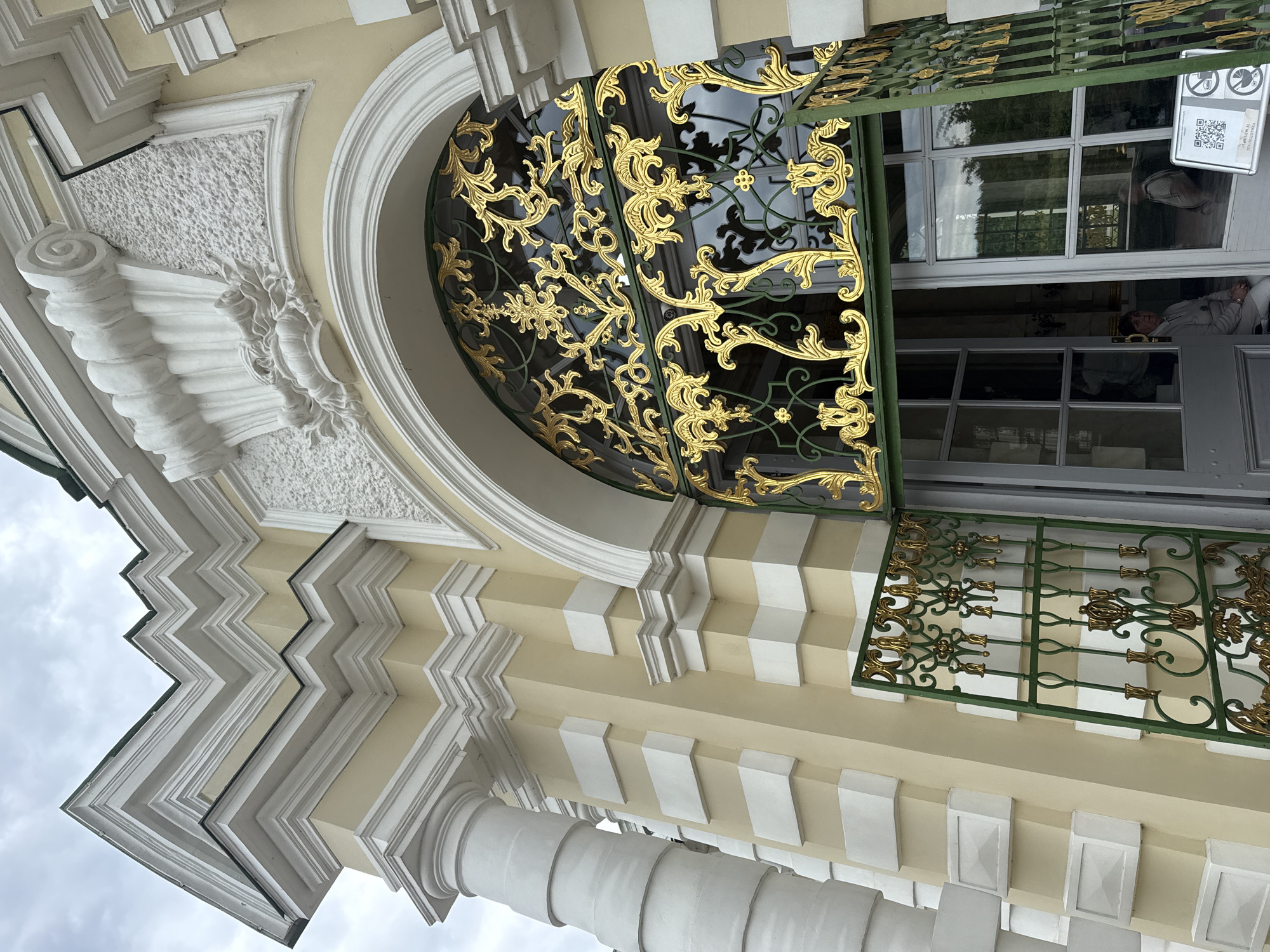 The image shows a low-angle view of a building facade in Moscow, Russia, specifically, the entrance to the Grand Palace in Tsaritsyno Park. The building features ornate Baroque-style architectural elements. There are white Doric columns. The facade is pale yellow with white decorative accents. A large arched window is adorned with an intricate gold lattice design. Green metal frames the arched window. The open door has a window with a QR code sign affixed to it. Inside the building, a person is reclining on a seat. The sky is overcast.