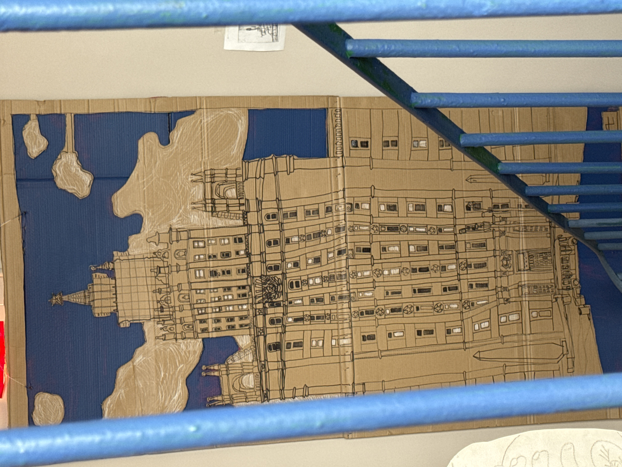 The image shows a drawing of the Seven Sisters skyscrapers in Moscow, Russia, on a piece of cardboard. The cardboard is rectangular and hangs on a white wall. The buildings are depicted in brown outlines with details showing windows and architectural elements. The area around the buildings is colored blue, suggesting water or the sky. The view of the drawing is partially obscured by blue metal bars, suggesting a railing or some kind of structure in the foreground.