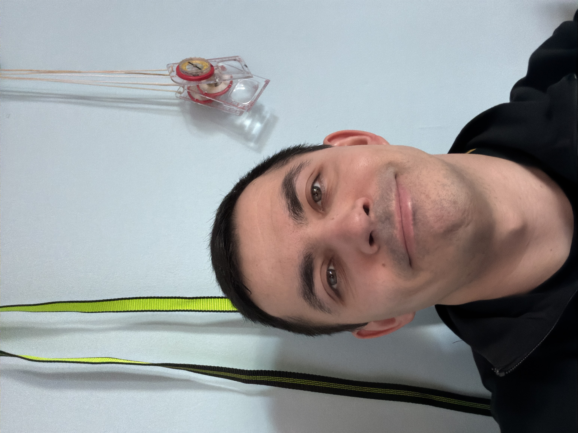 A selfie taken by a man with short, dark hair and brown eyes. He is wearing a black hoodie. In the top left corner of the image is a small, clear plastic device with red and white components and thin strings extending from it. Below the man, in the bottom left corner, is a yellow and black striped strap. The background is a pale blue wall. The location and city of Izhevsk, Russia, cannot be determined from the visual information provided.