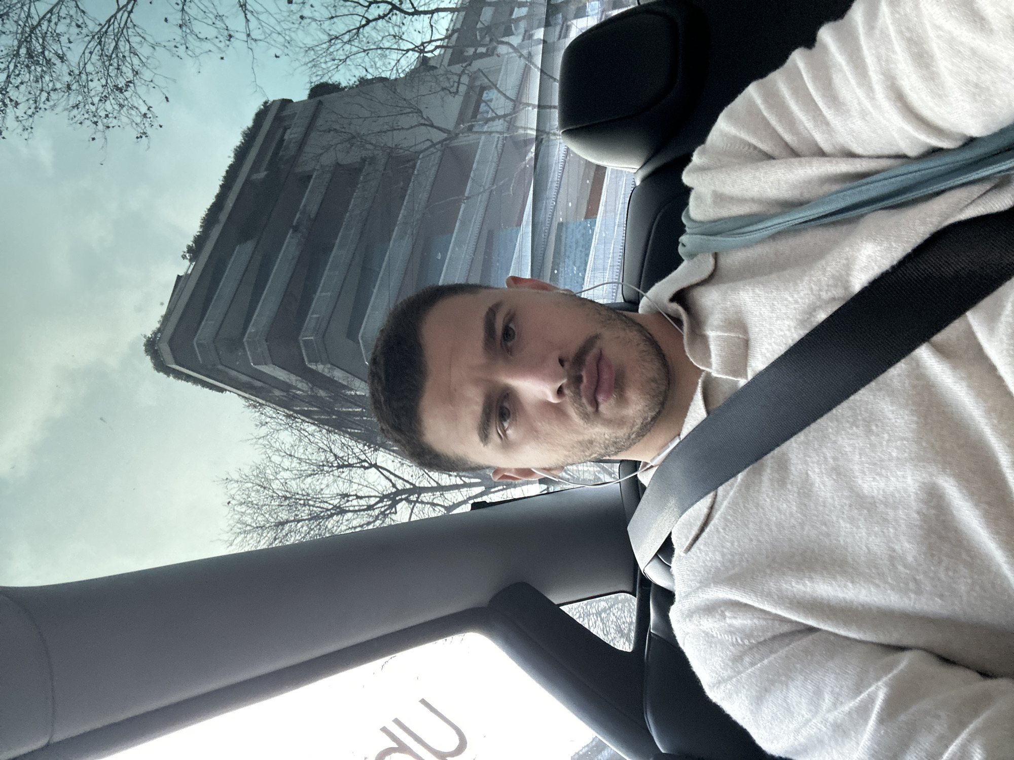 A male individual is depicted in a close-up, chest-up shot from within a vehicle, occupying the rear passenger seat. He wears a dark seatbelt across his chest and left shoulder. The subject has short, dark hair, a stubble beard, and a mustache. He is wearing a light-colored, possibly beige or off-white, long-sleeved polo-neck sweater and white wired earphones. A light blue strap, potentially from a bag, is visible over his left shoulder. He maintains direct eye contact with the camera, displaying a neutral expression.
Through the vehicle's windows, an urban environment under an overcast sky is visible. Bare deciduous trees with sparse brown leaves line the street. The most prominent external feature is a multi-story concrete building characterized by numerous recessed floors with balconies and extensive integrated greenery on its terraces and rooftop. This building is identified as one of the towers of the **Bosco Verticale (Vertical Forest)** complex, located in the Porta Nuova district of Milan, Italy. The perspective indicates the vehicle is situated on a street adjacent to this architectural landmark, such as Via Gaetano de Castillia or Via Confalonieri.
