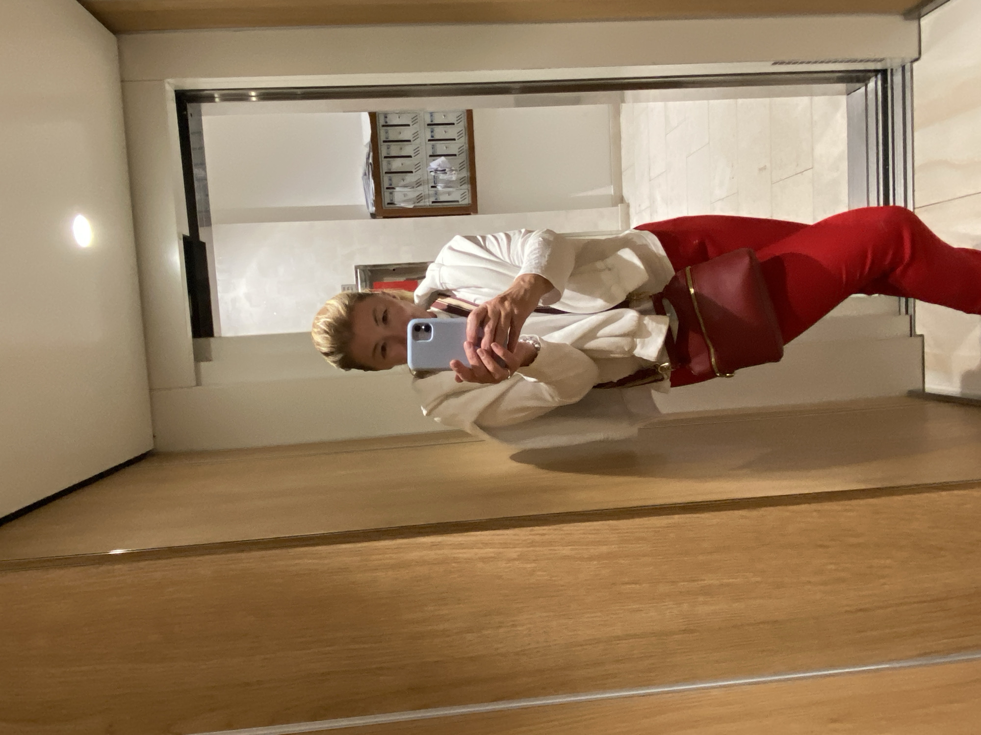 A mirror selfie captures a woman in a modern indoor setting, likely a building in Budva, Montenegro. The image, presented sideways, shows the woman standing upright before a large mirror, taking her photo. She has blonde hair tied back and is wearing a white long-sleeved shirt and bright red trousers. A red shoulder bag with gold details is worn across her body. She holds a light blue smartphone, taking the picture, and her gaze is directed towards the phone's camera in the reflection.
The mirror's reflection reveals the interior space behind the woman. To the left, a white wall features a circular, glowing light fixture. In the background, a framed unit resembling a wall of mailboxes or a directory, filled with papers, is mounted on a light-colored wall. The floor visible in the reflection is light-toned, possibly tiled. A dark, vertical element, potentially a door frame or elevator edge, is visible on the right.
The woman stands on a polished wooden floor, which constitutes the foreground of the image outside the mirror's frame. The environment suggests a clean, contemporary hallway, lobby, or common area within a building, utilizing artificial lighting. No discernible text is visible in the image.