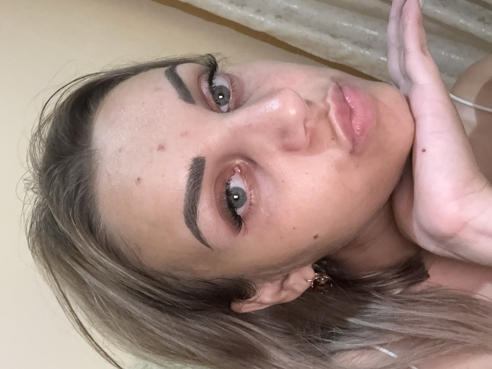 A close-up, medium shot shows a woman with blonde hair and light skin resting her head on her hand, looking towards the camera with pursed lips. Her eyes are open, and she is wearing false eyelashes. There is a small red blemish on her forehead and a mole on her cheek. She is wearing a white strap and a gold earring. The background appears to be a beige curtain. The image quality is poor, with visible noise and blur. The location in Kholmsk, Russia is not discernible from the image.