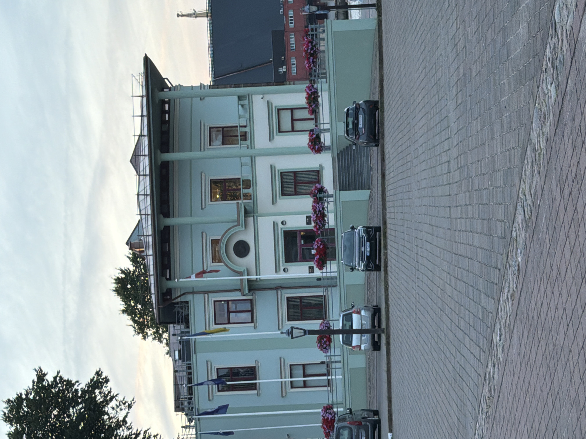 The image shows a light blue building with a flat roof and classical architectural elements, located in Ventspils, Latvia. The building has multiple windows with dark red frames and decorative flower boxes. Several flags are displayed on poles in front of the building, including the Ukrainian flag and what appears to be the Danish flag. A paved area in front of the building has four cars parked. The environment suggests a public space, possibly a municipal building or a cultural institution, adjacent to a waterfront, indicated by the presence of a boat mast in the background. The sky is overcast.
