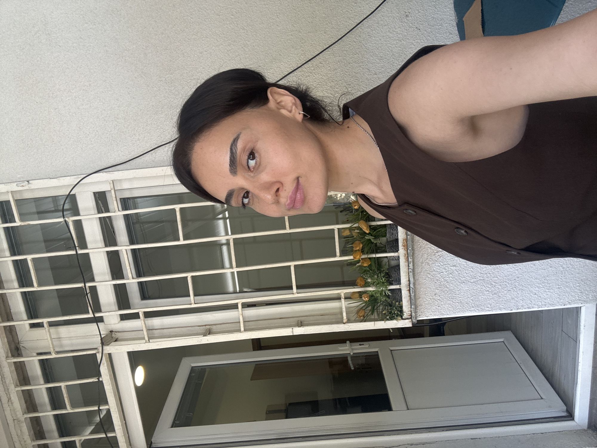 A young woman with dark hair pulled back and light skin is posing for a selfie outdoors. She is wearing a sleeveless brown top. Her expression is neutral, and she is looking directly at the camera. Behind her, a white door with glass panels is open, revealing a glimpse of an interior space. To the right of the door is a white, barred window with a planter box containing artificial yellow flowers. The background appears to be a textured white wall. The lighting suggests it is daytime. The location is identified as Tbilisi, Georgia.