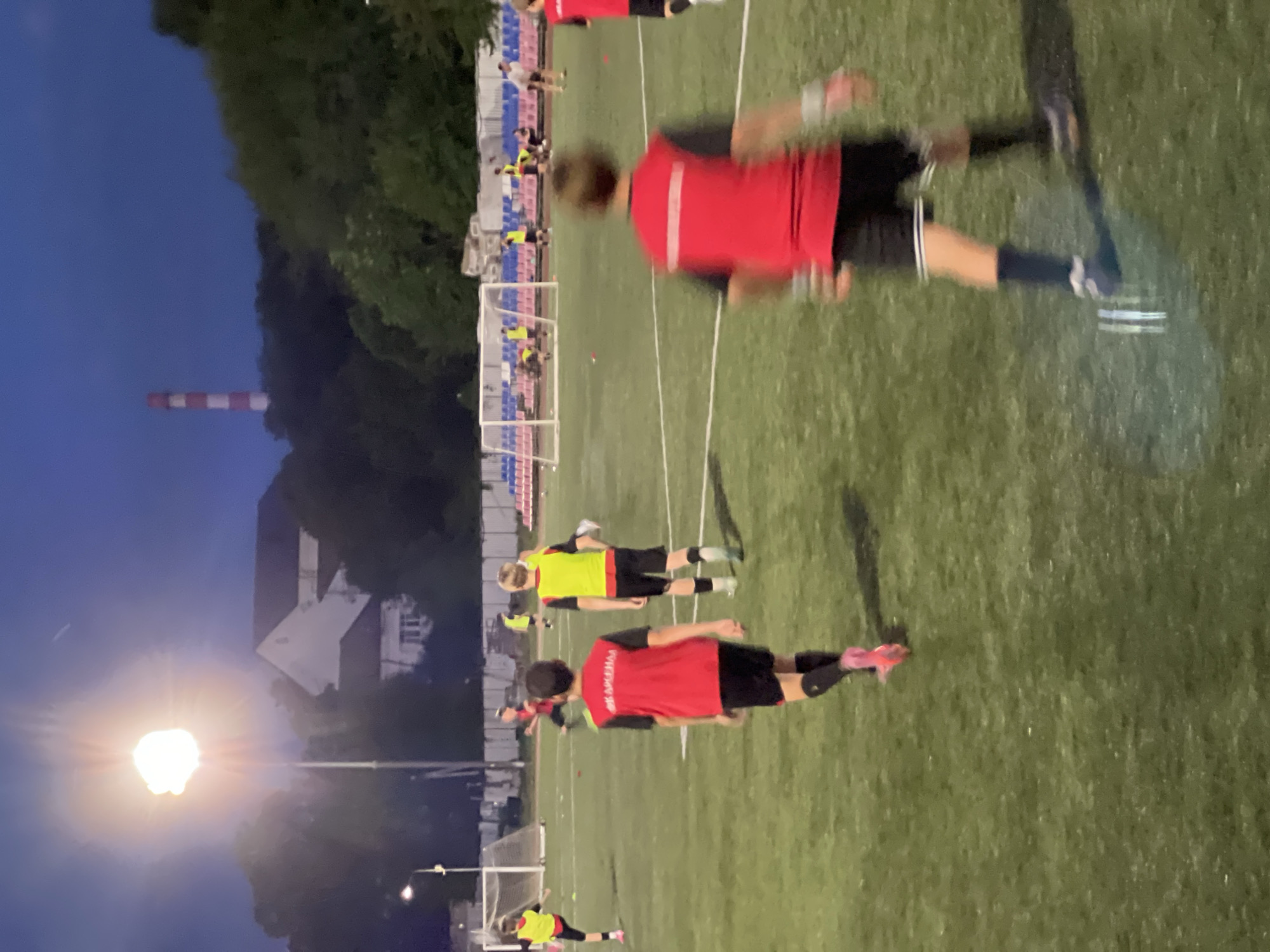 The image depicts a nighttime outdoor soccer practice session on an artificial turf field. Several young male individuals, likely pre-teens or early teens, are present, clad in red and black soccer uniforms or practice vests over their uniforms. Some are actively engaged in drills or practice, with one individual in the foreground captured in motion, suggesting a dynamic action shot. The field is illuminated by bright artificial lights, casting distinct shadows and creating a well-lit playing surface. In the background, a sports stadium with seating and goalposts is visible, along with trees and buildings, including a tall, red-and-white striped industrial smokestack which may indicate the proximity to industrial areas. The overall setting suggests a community or youth sports facility. Krasnodar, Russia, features numerous such sports complexes, often equipped with artificial turf fields for year-round use, and is known for its mild climate allowing for outdoor sports activities into the evening. The presence of the smokestack is a common sight in industrial regions of Russia.