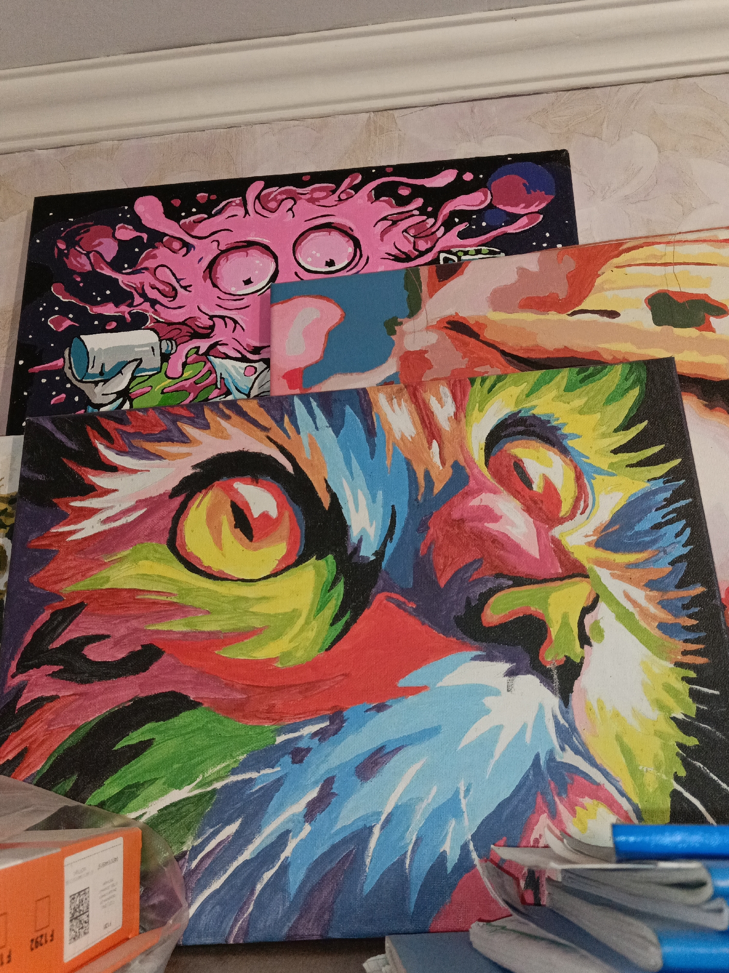 The image shows three canvases stacked on top of each other, leaning against a textured wall. The top canvas depicts a cartoonish pink creature with large, surprised eyes against a dark background with white speckles and a blue and pink planet. A white and teal cylindrical object is being held by the creature.
The middle canvas, partially visible, shows a stylized, abstract landscape with bold colors of orange, yellow, pink, and blue, with some green elements.
The bottom and largest canvas features a vibrant, close-up portrait of a cat's face rendered in a bold, colorful, almost psychedelic style. The cat's eyes, a striking yellow with black pupils, are the central focus. The fur is painted with broad strokes of various colors, including red, blue, green, yellow, white, black, and purple, creating a dynamic and energetic impression.
In the foreground, at the bottom left, is a partially visible orange box with a white label. The label contains a barcode and some text. Visible text on the orange box includes "F1292". Also visible are several blue pens and stacks of paper or other flat items resting on top of the bottom canvas.
The lighting suggests an indoor setting, with no indication of time of day or weather. The overall impression is that of a collection of artistic works, possibly paintings completed or in progress. The location context provided is Shymkent, Kazakhstan, but there are no specific visual cues within the image to confirm this.