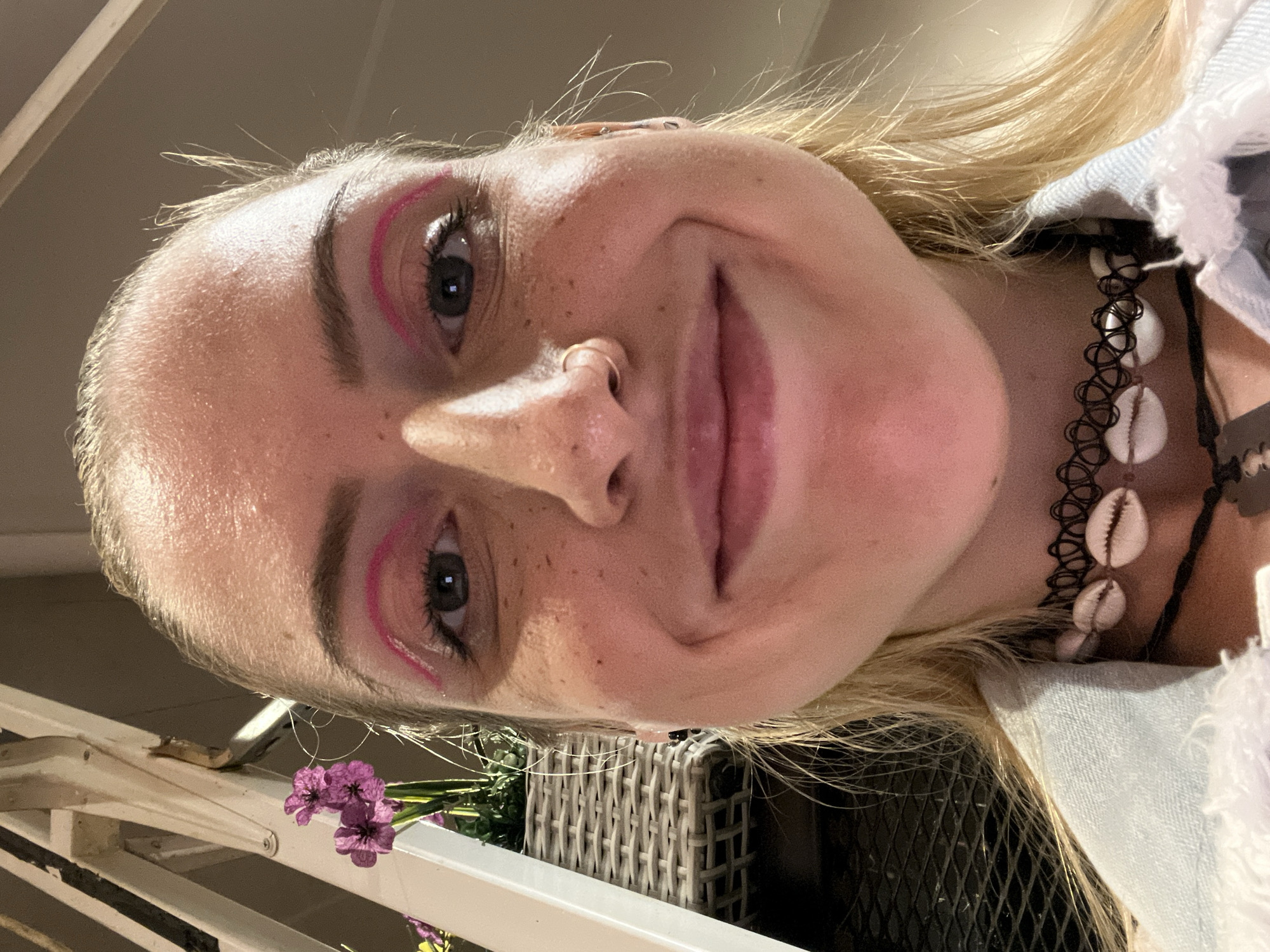 A close-up, slightly angled, selfie-style shot of a blonde-haired woman with freckles. She has a nose ring and is wearing pink eyeliner. A black choker with white shells is visible around her neck. In the foreground are purple flowers and a white-painted railing. A wicker basket is also visible in the background. The location is not discernible in the image. There is no information within the image that allows one to identify the location as Plovdiv, Bulgaria.