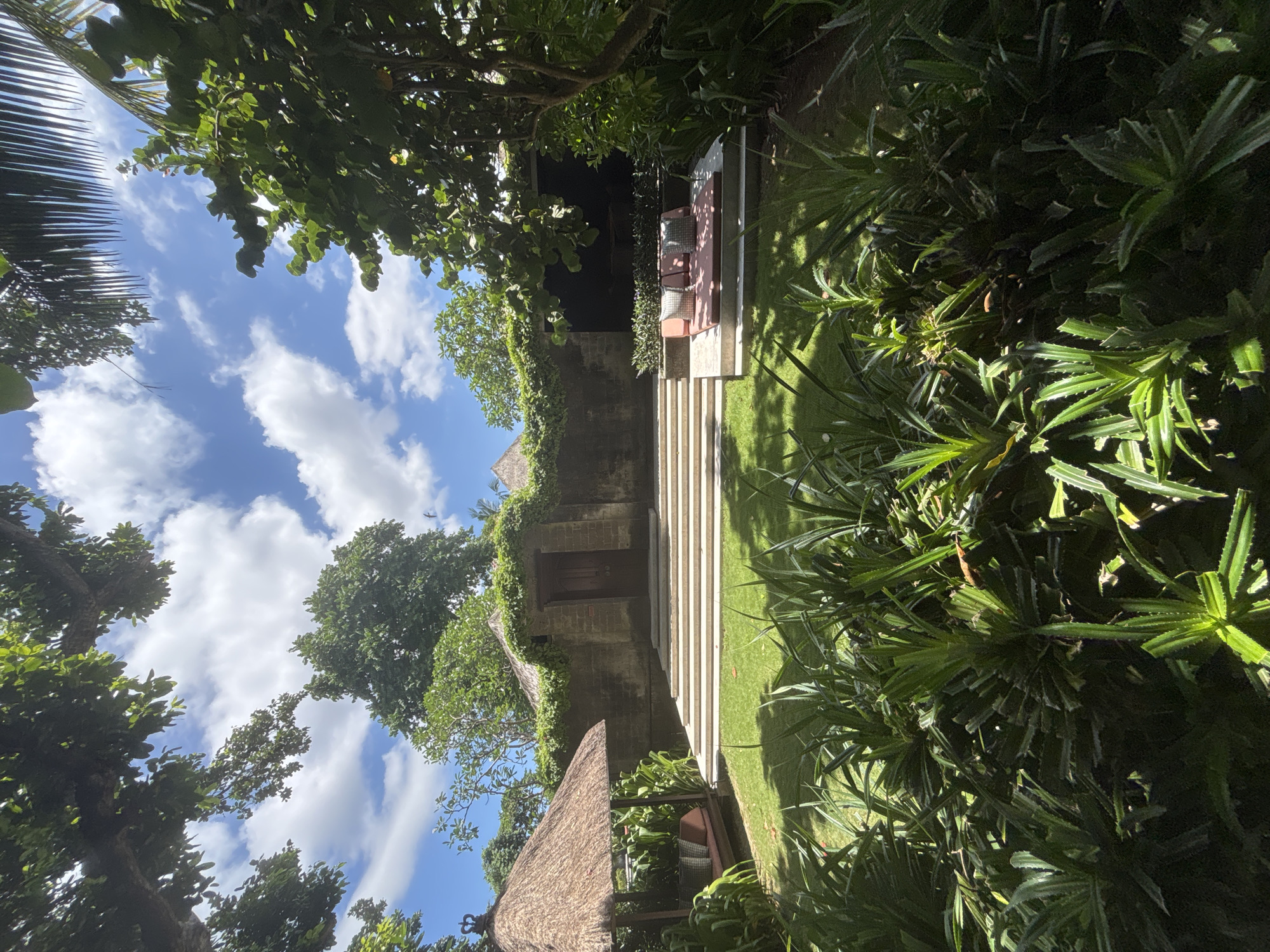 The image captures a tranquil outdoor setting, likely a garden or courtyard in Denpasar, Indonesia. A stone structure with a wooden door is partially covered in greenery and accessible by a set of stone steps flanked by grass. A separate thatched-roof structure provides shade to an outdoor lounge area with a cushioned seating arrangement. Lush tropical foliage frames the scene, obscuring parts of the sky with visible clouds. The orientation of the image is rotated counter-clockwise.