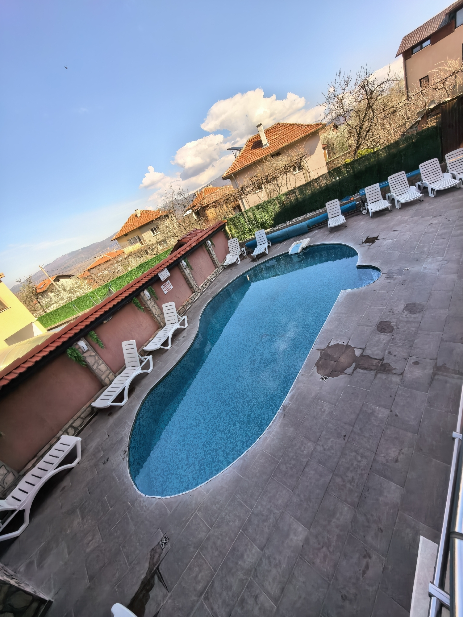 The image captures a swimming pool area in an unidentified city. The pool is surrounded by gray stone tiles. White lounge chairs are positioned around the pool, some along a light brown stone wall topped with green foliage. Buildings with red-tiled roofs are visible in the background, set against a backdrop of blue sky with fluffy white clouds. The scene appears deserted, with no people present.