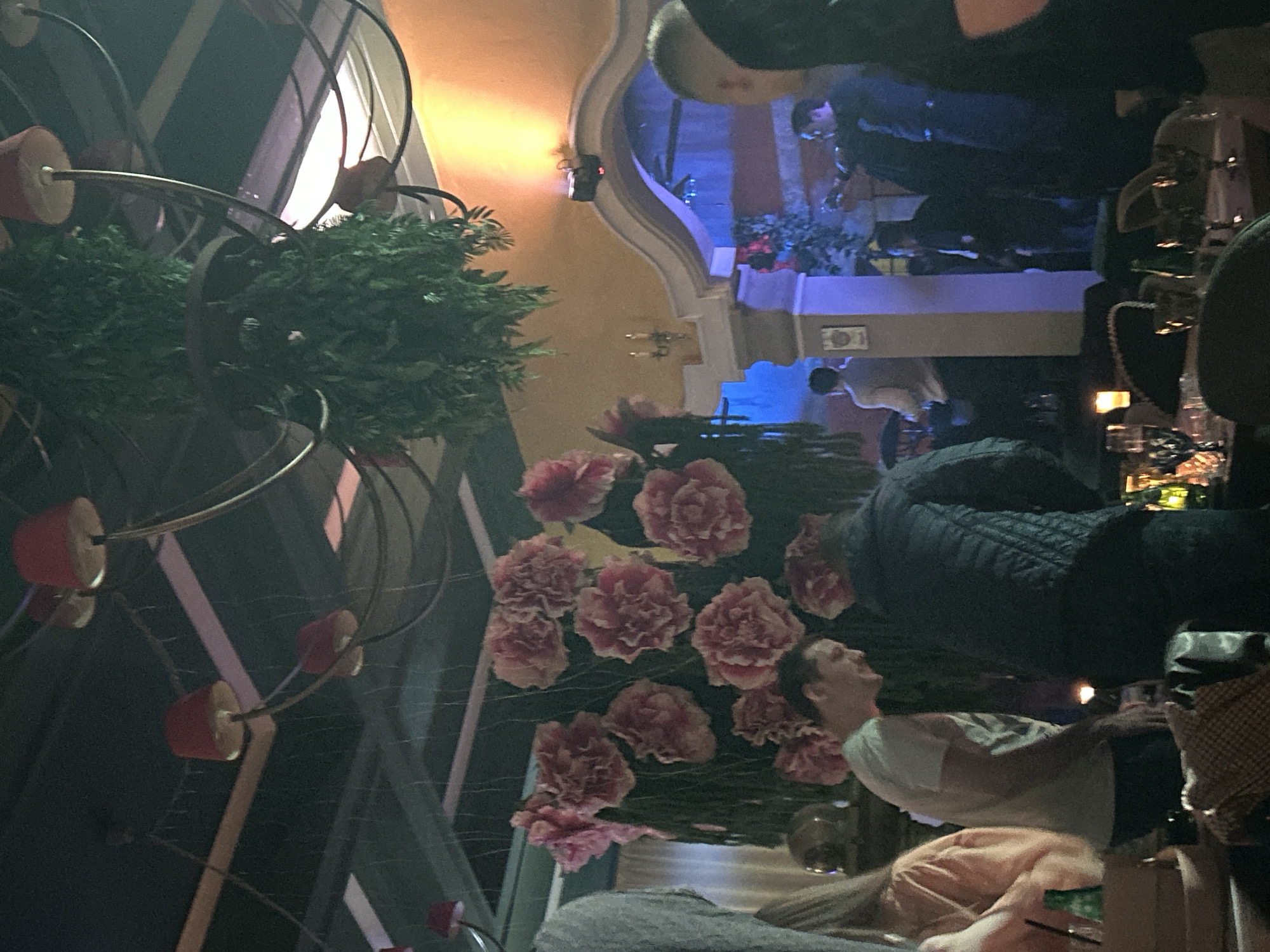 The image shows an interior setting, likely a bar or restaurant in Sofia, Bulgaria. People are seated at tables; some are interacting, while others are occupied with drinks and conversation. The environment is illuminated by chandeliers with exposed pink bulbs. In the background, an archway and a blue-lit area suggest a different space within the establishment. Decorative elements such as flower arrangements and greenery contribute to the interior's ambiance. The tables are set for service, with glassware and other dining essentials. The overall atmosphere is dimly lit, creating a mood conducive to social interaction.