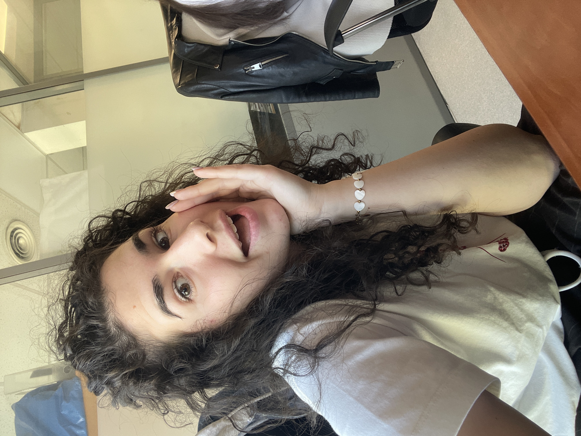 A close-up, portrait-style photo captures a young woman with dark, curly hair and light brown eyes, looking directly at the camera. Her head is tilted, and her left hand is raised to her cheek, displaying long, lightly polished fingernails. Her mouth is open in an expressive manner, suggesting surprise or a playful laugh. She is wearing a simple white t-shirt. On her left wrist, she sports a delicate bracelet featuring several heart-shaped, white mother-of-pearl beads interspersed with gold spacers.
The background shows an indoor setting, likely a modern building. To the woman's left, a light-colored, possibly frosted glass or panel partition is visible, with horizontal lines. Above this, a circular air vent can be seen on a white ceiling. A blue object, possibly a bag, is partially visible in the bottom left corner. Behind her, a black leather jacket with a prominent silver zipper and buckle details is draped over what appears to be a chair or piece of furniture. A white ribbed sweater is visible underneath the jacket. On the right side of the image, an orange-brown surface, possibly a wall or table, fills the frame. The lighting suggests it is daytime, with bright, even illumination. No specific location cues for Sofia, Bulgaria are visible in the image itself, though it appears to be a public or professional indoor environment. No visible text is present.