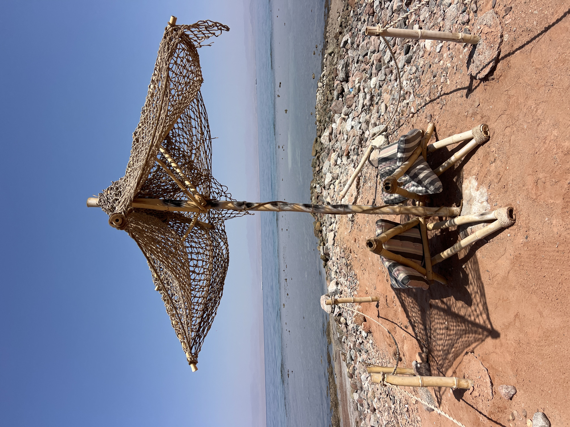 Two woven bamboo chairs with striped cushions sit on reddish-brown sand beneath a large, netting-covered umbrella. The umbrella is supported by a bamboo pole wrapped with what appears to be rope or twine. The chairs and umbrella cast shadows on the sandy ground. Bamboo poles with rope barriers delineate the seating area. Beyond the sand, a rocky shore meets the calm, blue sea. In the distance, mountains are visible under a clear, bright blue sky. The scene is located at a beach in Nuweiba, Egypt.