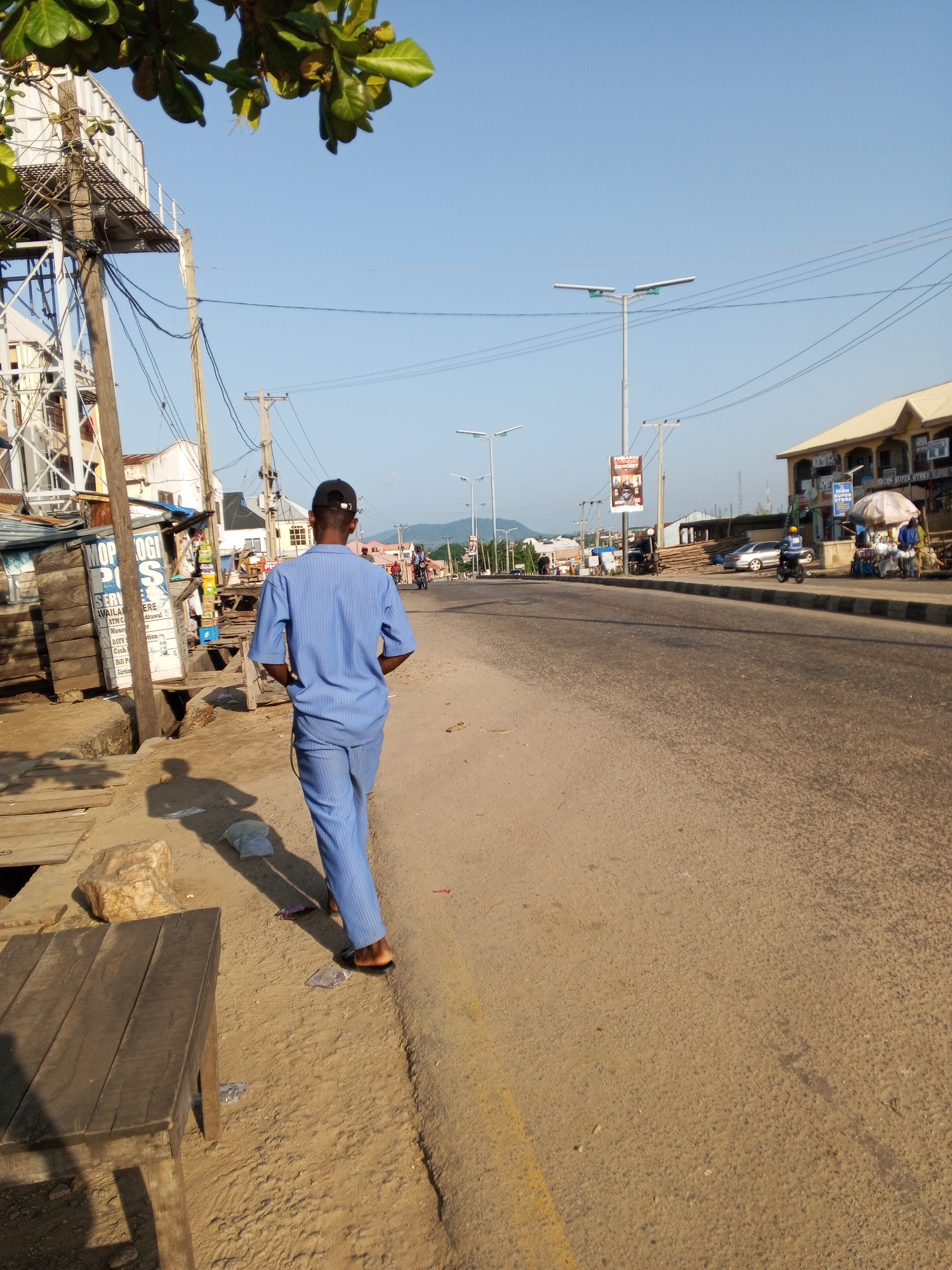 This media file captures a sunny daytime scene in Lokoja, Nigeria, likely along a moderately busy street. The foreground is dominated by a man walking away from the camera, dressed in a light blue, matching two-piece outfit and a black baseball cap. He has his hands in his pockets, suggesting a relaxed or casual demeanor. Next to him, on the left side of the image, is a wooden bench. The ground is sandy and uneven, with some debris and a large rock.
The background reveals a street with a few cars, motorcycles, and other pedestrians. Buildings line the right side of the street, including one with a sign reading "ICON SUPER STORE." Streetlights and utility poles with numerous wires are also prominent. The overall setting appears to be a commercial or residential area, with a mix of structures and open space. The clear blue sky indicates fair weather. The presence of a street vendor with an umbrella adds to the impression of a lively, everyday scene in this Nigerian city. Visible text includes "MOP," "SERV," "ICON SUPER STORE," and other partial words that are difficult to decipher completely.
