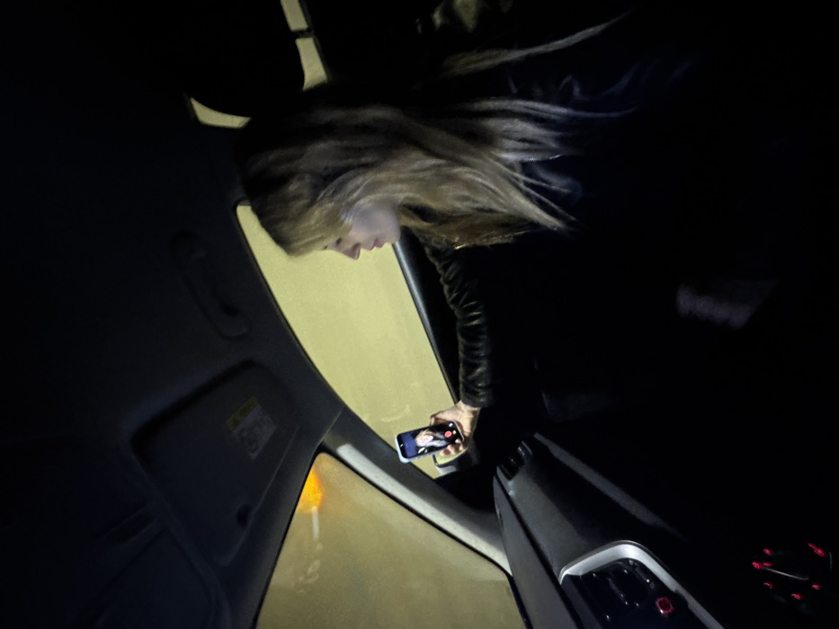 The image depicts the interior of a vehicle, likely positioned within the Sozina Tunnel in Montenegro, indicated by the pervasive uniform yellowish-green light illuminating the exterior visible through the windows. An orange light source is also discernible through the upper left portion of the windshield. A female individual, with long, light-colored hair, is seated in the front passenger seat, wearing a dark jacket. She holds a smartphone in her extended right hand, actively engaged in a video call, with a person's face visible on the illuminated screen. Her profile is visible, and her gaze is directed forward, seemingly towards the phone or the scene outside. The car's dashboard features air vents and control buttons illuminated with red light.