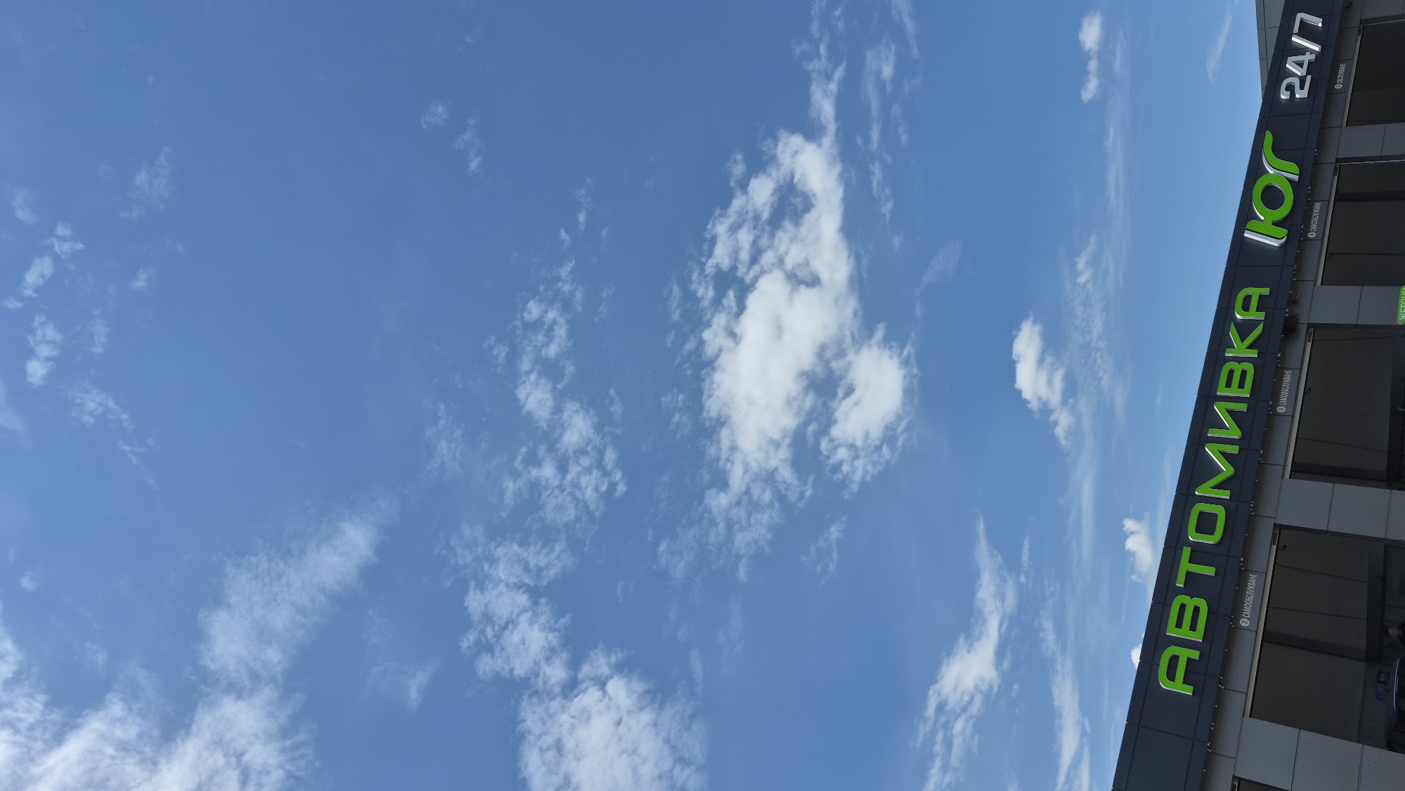 An outdoor, eye-level, vertical medium shot shows the bright blue sky filled with white, wispy clouds above a dark gray building with large green lettering that reads "АВТОМИВКА ЮГ 24/7". The word "АВТОМИВКА" is written in large, capital, green, sans-serif letters, followed by "ЮГ" in a similar style, and "24/7" in white, capital, sans-serif letters. Below the main sign, smaller text in white reads "САМООБСЛУЖИВАНИЕ" in two places. The building appears to be a car wash. The sky occupies roughly two-thirds of the frame, with the building filling the bottom third. The lighting suggests it is daytime, with sunlight illuminating the clouds and the building. No people are clearly visible within the frame.