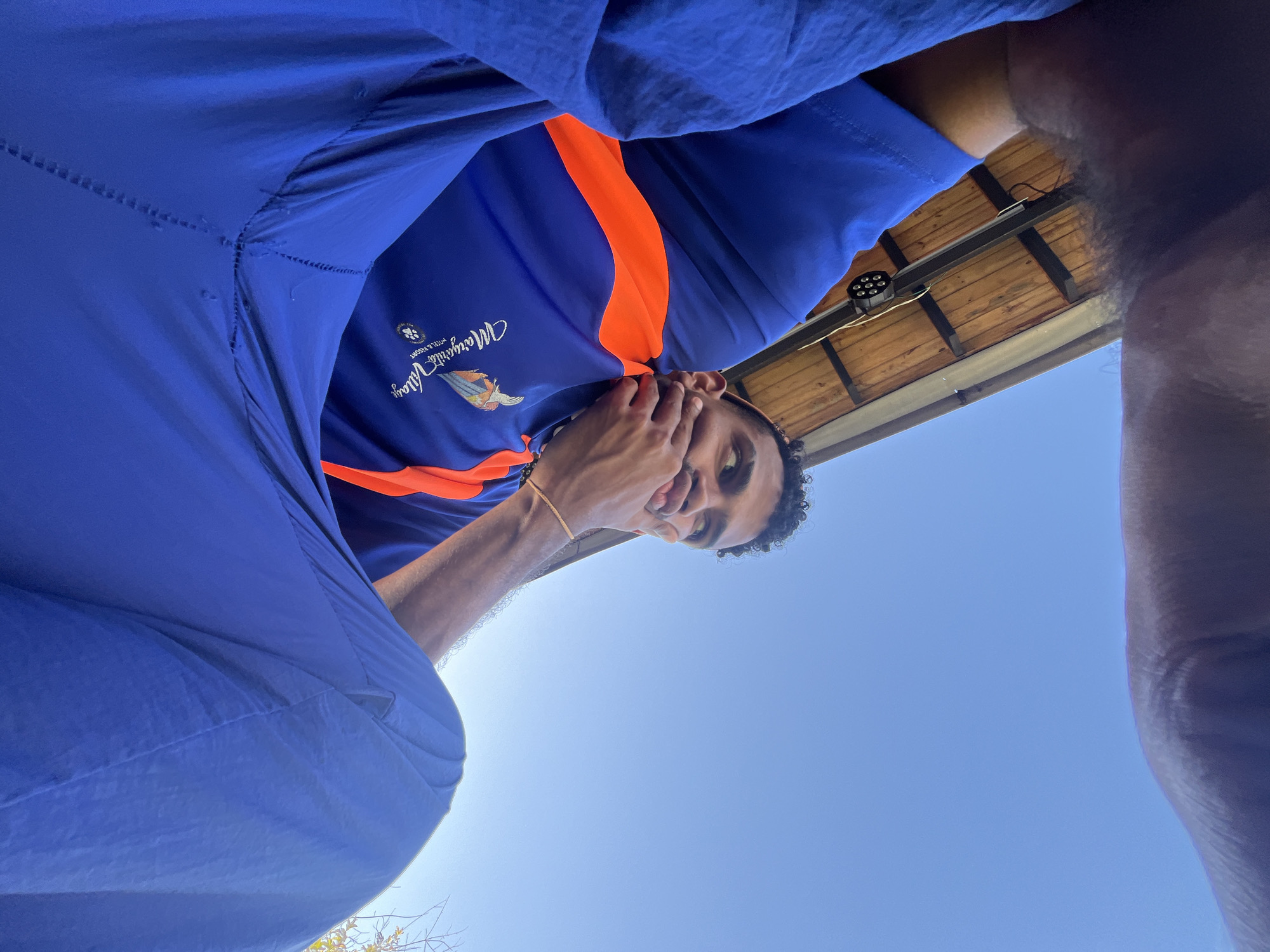 A man with curly dark hair is looking down at the camera, partially covering his mouth with his left hand. He is wearing a blue shirt with an orange stripe across the chest, with a logo that reads "Margarita's Village Hotel & Resort". The location appears to be outdoors with a clear blue sky in the background. There is a section of a wooden roof with light fixtures visible above his head. The overall perspective suggests the camera is positioned below the man, looking up at him. Given the logo on the shirt, the location is most likely at or near the Margarita's Village Hotel & Resort in Porlamar, Venezuela.