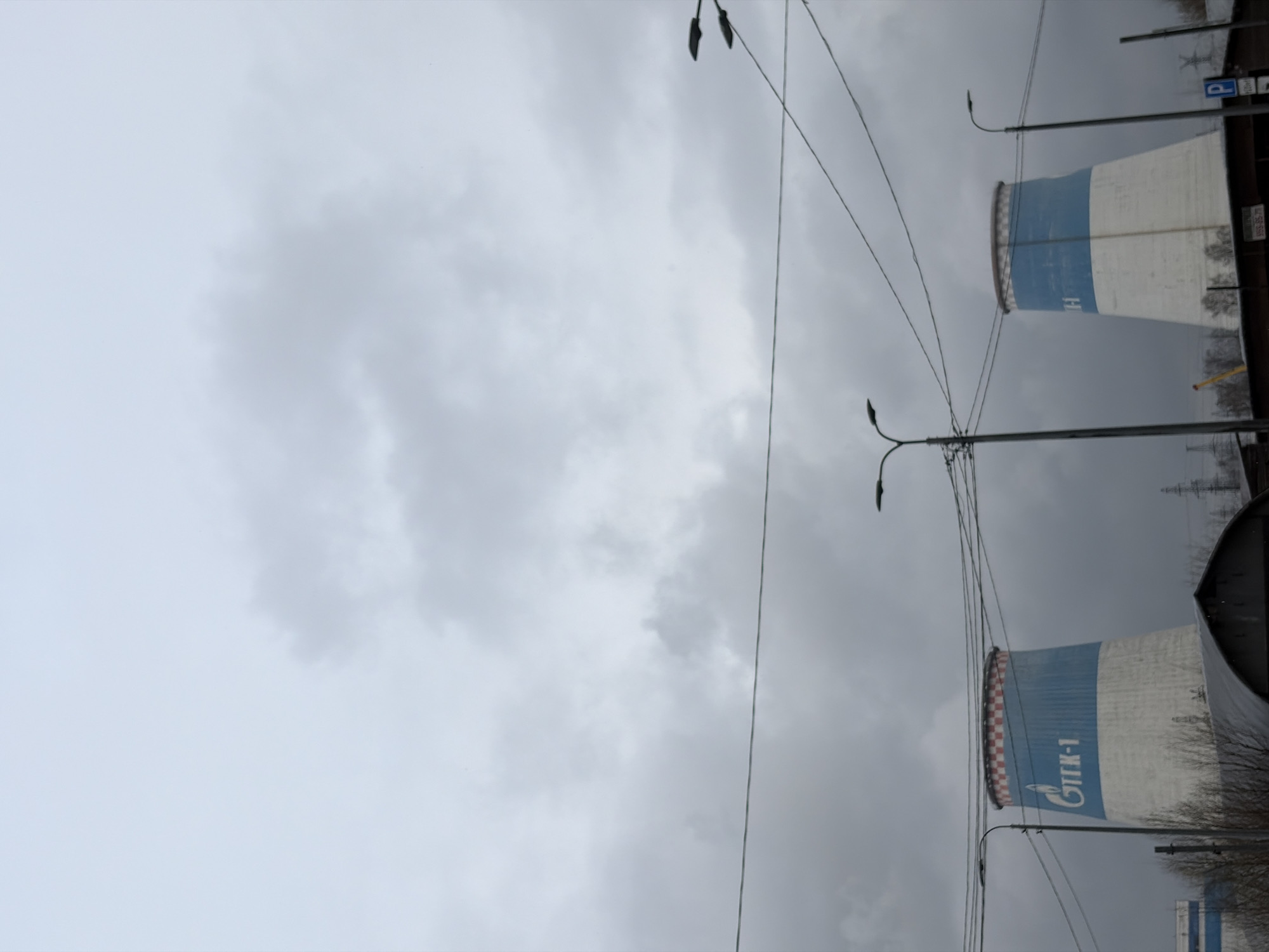 The image shows a view in Saint Petersburg, Russia, featuring two large cooling towers. The cooling towers have a blue and white paint scheme and bear the logos of "Gazprom" and the inscription "CHP-1". Power lines and streetlights are visible in the foreground, partially obscuring the view of the cooling towers. The sky is overcast and gray. The tops of bare trees are visible along the bottom.