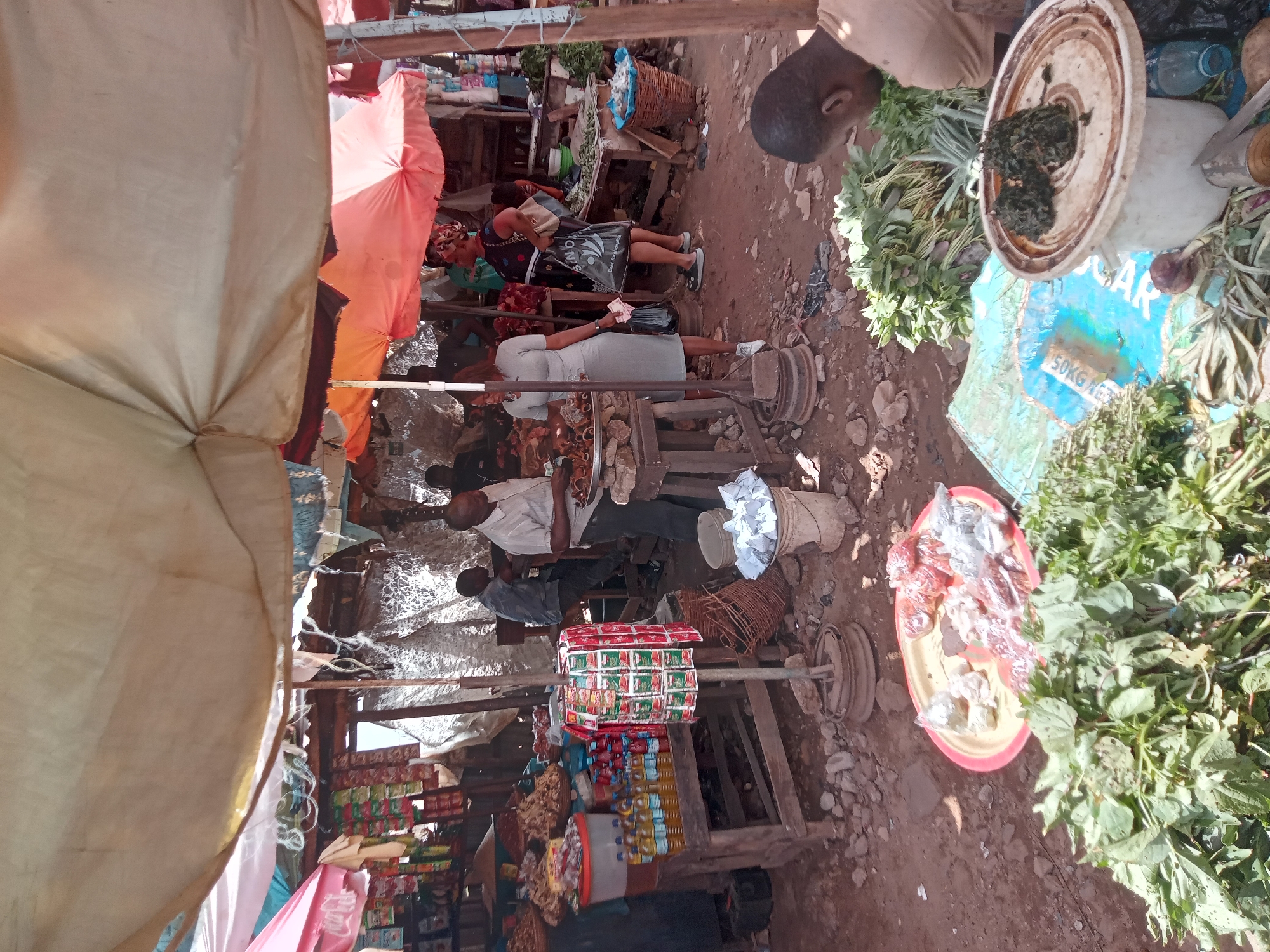 The image shows a bustling market scene in Sabon Tasha, Nigeria. Several people are present, including vendors and customers. One woman is paying a vendor. There are stalls selling various goods such as vegetables, snacks, canned items, and cooking oil. The stalls are makeshift structures, with tarps and umbrellas providing shade. The ground is dusty and uneven. A scale is visible near a vendor and customer, suggesting items are being weighed for purchase.