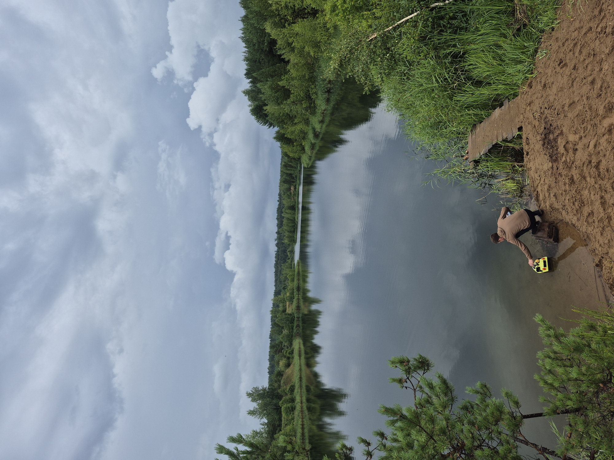 In the city of Novyi, Russia, a person is by the edge of a calm lake, partially obscured by trees and reeds. The person is crouching and appears to be interacting with a yellow toy boat in the shallow water. A small wooden pier extends from the sandy bank towards the lake. The sky is overcast with dramatic clouds, and the surrounding landscape is densely forested, with reflections of the trees visible on the water's surface. The overall scene depicts a serene, natural environment on a cloudy day.