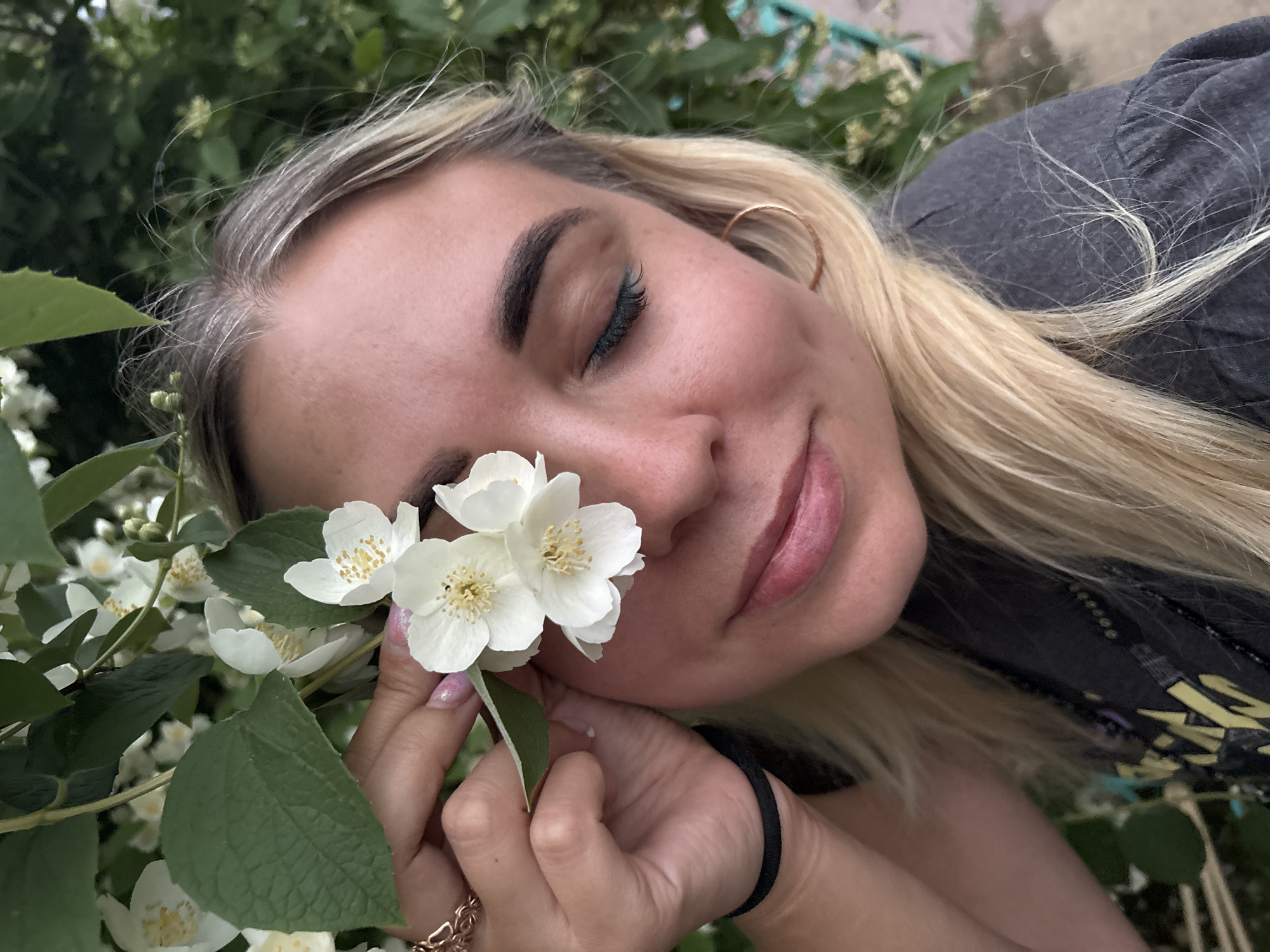 A young woman with blonde hair is in close-up, with her eyes closed and a soft smile. She is holding several white jasmine flowers to the right side of her face, obscuring part of her eye and forehead. She is wearing a grey t-shirt and has a black elastic band on her left wrist. Her fingernails are painted with a light pink glitter polish. A gold ring with a floral design is visible on her left index finger, and a gold hoop earring is worn on her right ear. The background is filled with green leaves and more white flowers, suggesting an outdoor setting. The lighting is soft and natural. The scene appears to be in Chaikovsky, Russia, given the context of the request.