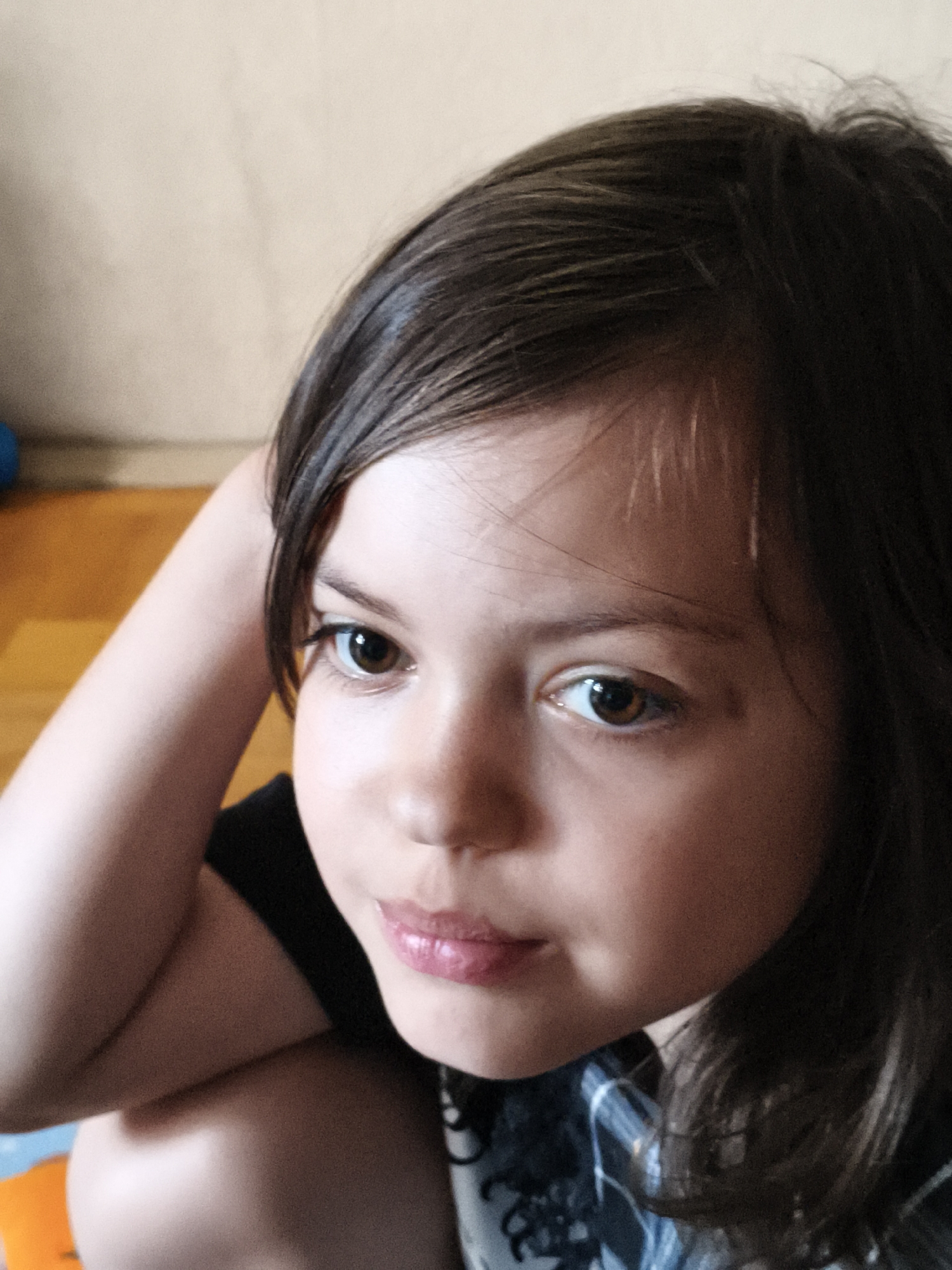 This is a close-up, head-and-shoulders portrait of a young girl. She has dark brown hair that falls across her forehead and frames her face. Her eyes are large and brown, with a thoughtful or perhaps slightly sad expression. Her lips are slightly parted, with a hint of pink lipstick or gloss. One of her arms is bent and raised behind her head, with her hand resting near her temple. She is wearing a dark shirt, possibly with a pattern that is partially visible. The background is blurred, but it appears to be an indoor setting with a light-colored wall and a wooden floor. There are hints of blue and orange objects in the far background, suggesting a play area or living space. The lighting suggests it is daytime, possibly indoors with natural light coming from the side. The overall mood is intimate and candid, capturing a moment of quiet reflection.