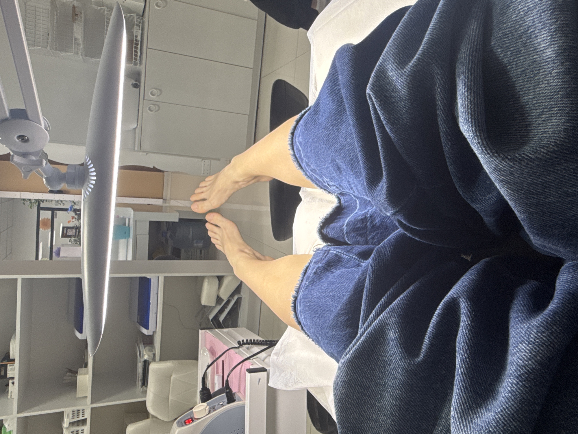 A person wearing denim shorts is lying on a white treatment table in a room. The person's feet are visible. Various pieces of equipment and shelving are present. The location is Batumi, Georgia.