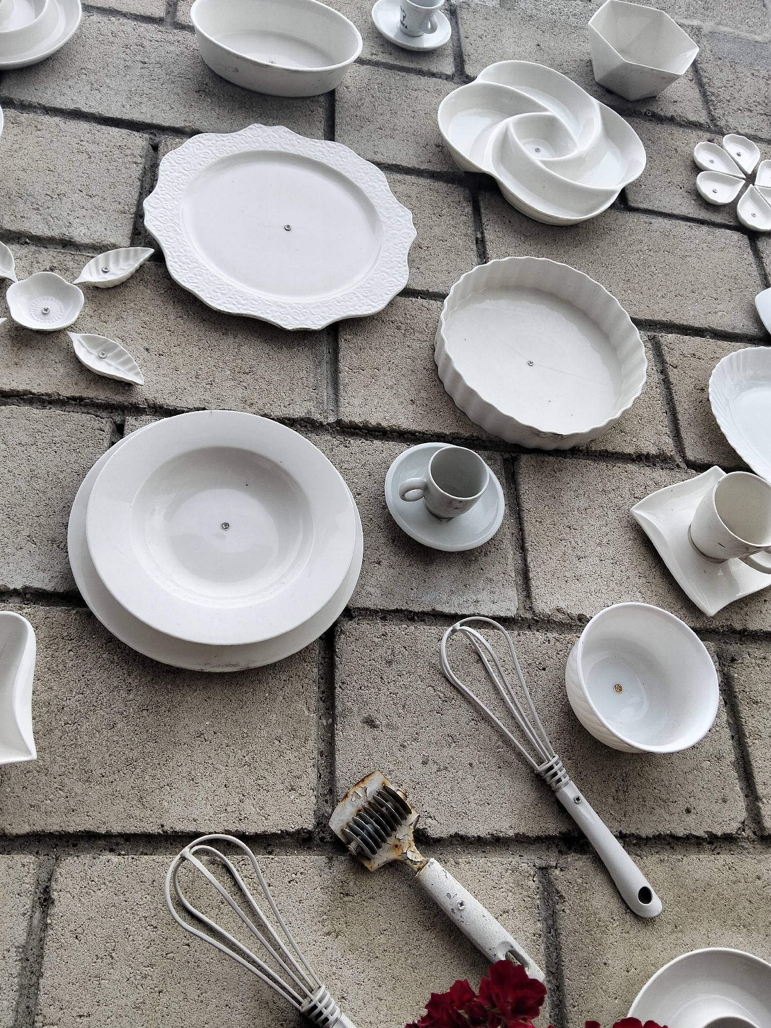 The image displays a collection of white ceramic kitchenware and utensils mounted on a grey brick wall in Peresecina, Moldova. The items include various plates, bowls, cups, saucers, a whisk, and a meat tenderizer. Some of the plates are decorative with embossed patterns, while others are plain and functional. The items are arranged at different angles and heights, creating a visually interesting composition. The brick wall appears weathered, with visible grout lines and texture. The lighting suggests an outdoor setting, likely during the day. There are no people or animals present in the scene.