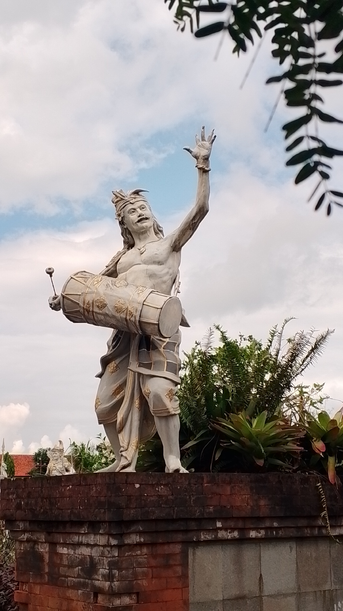 The media file depicts a stone statue of a man in traditional clothing, holding a drum. The statue is positioned on top of a brick structure, surrounded by lush green foliage. The man is depicted with a handlebar mustache and a headdress, and his right arm is raised in the air. The drum, adorned with ornate patterns, is held horizontally in his left arm.
The setting appears to be outdoors, with a cloudy sky visible in the background. The lighting suggests it is daytime, possibly overcast due to the diffused light. The statue's pose and the drum suggest a performance or a ceremonial scene, perhaps depicting a musician or dancer.
Notable details include the intricate patterns on the drum and the man's clothing, as well as the texture of the brickwork beneath the statue. The presence of greenery indicates a possibly tropical or garden-like environment. No visible text is present in the image.