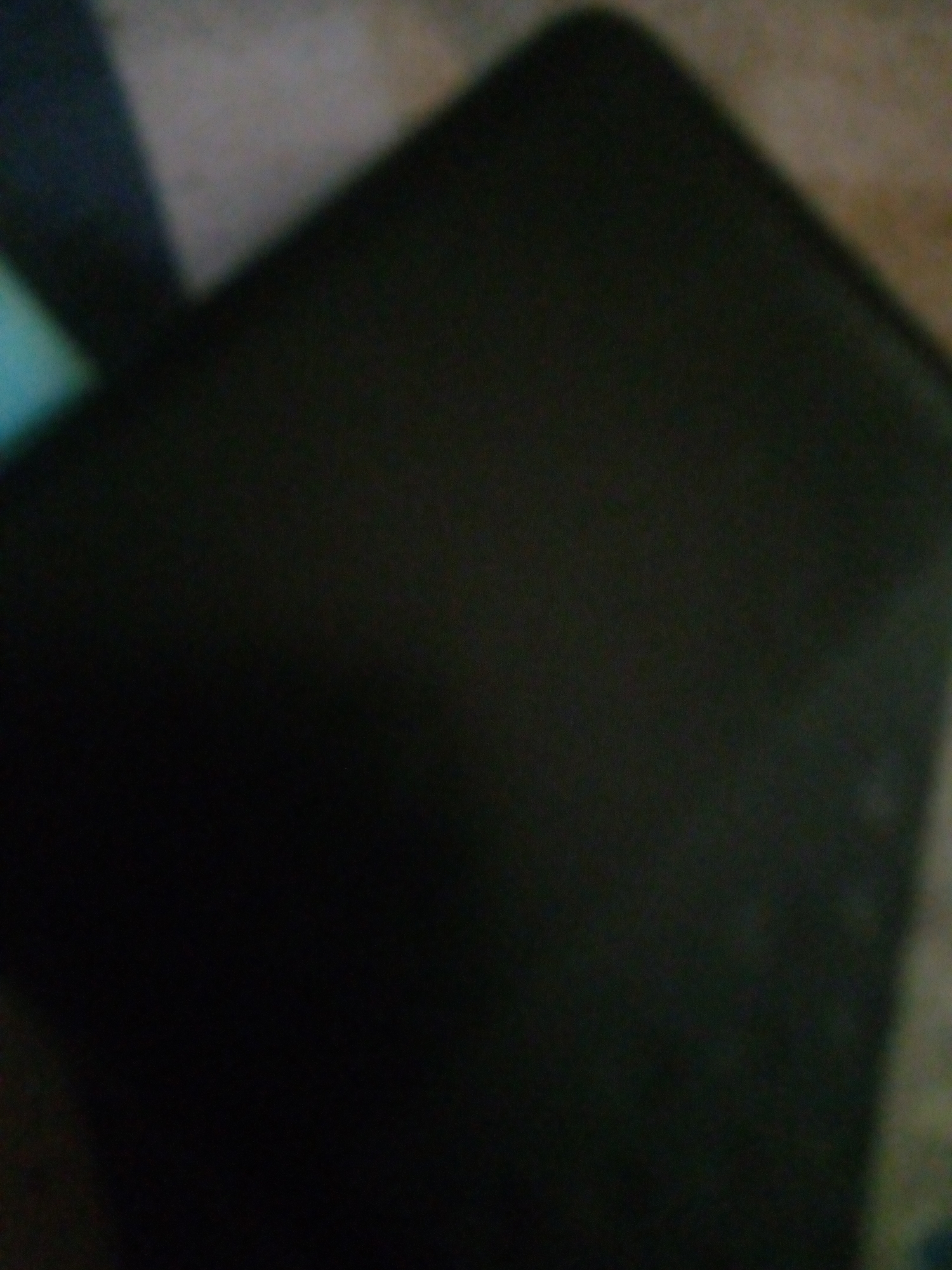 A dimly lit, blurry image shows a dark, rectangular object, possibly a book or a laptop, resting on a textured surface. The surface appears to be a combination of a rug or carpet and a lighter, possibly wooden or tiled area. The lighting is poor, with shadows obscuring fine details. The overall impression is one of an indoor setting, potentially a room in Jalingo, Nigeria, but there are no distinct landmarks or identifying features to confirm the location. The image is devoid of any people, actions, or interactions.