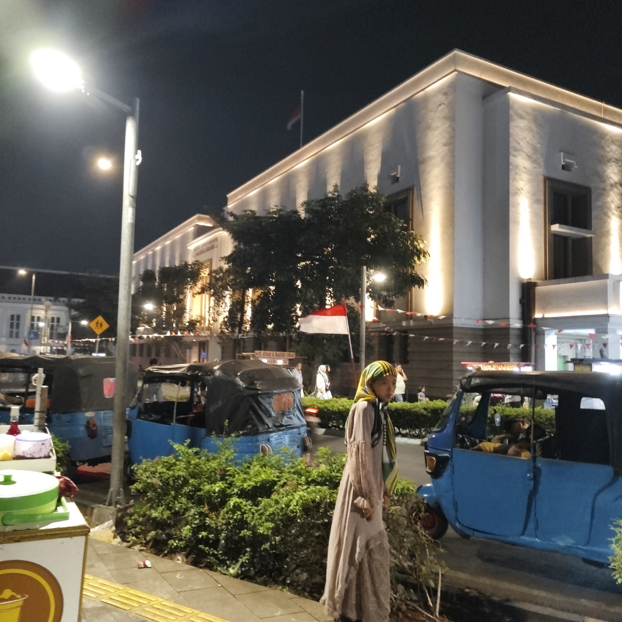 This is a nighttime, medium-distance shot of a street scene in West Jakarta, Indonesia. The foreground features a woman in a long, light-colored dress and a headscarf walking past a row of parked three-wheeled vehicles, likely Bajaj. One blue Bajaj is prominently featured on the right side of the frame, and its driver is visible inside. To the left of the Bajajs, there is a vendor cart with various containers, one of which is green and lidded. A lush green hedge runs along the sidewalk. In the background, a large, modern building with illuminated architectural lighting is visible. A flag, appearing to be the Indonesian flag with its red and white horizontal bands, flies from a pole on this building. Further in the background, other buildings and streetlights can be observed. The overall impression is a vibrant street scene at night, with a mix of traditional and modern elements.