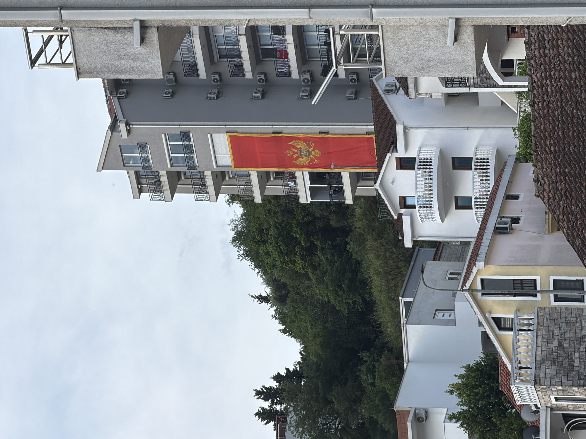 The image shows a view of buildings in Budva, Montenegro. A gray apartment building prominently displays the flag of Montenegro. Balconies are a common feature on the buildings. Lush green trees fill the space between the buildings. The sky is overcast.