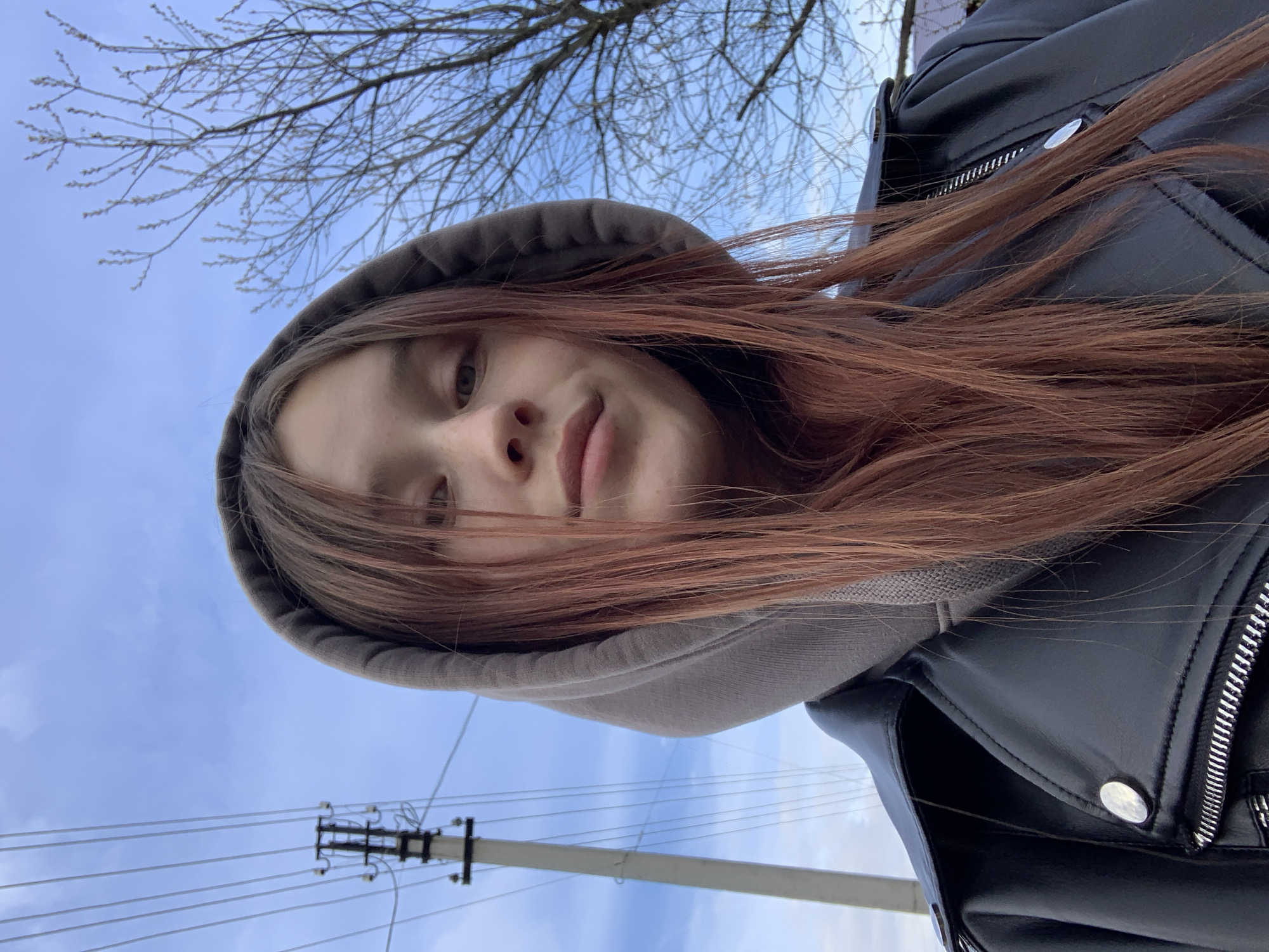 A photograph taken from a low angle, possibly by a person looking up, captures a young individual with reddish-brown hair wearing a gray hoodie underneath a black leather jacket. The individual's face is partially obscured by strands of hair falling across the forehead.
The backdrop reveals a light blue sky with sparse, bare tree branches at the upper left. Overhead power lines and a utility pole are visible against the sky. Based on the wires and pole structure, it is likely the photograph was taken in the vicinity of a populated area. Given the location, the location might be the city of Фоки, Russia.