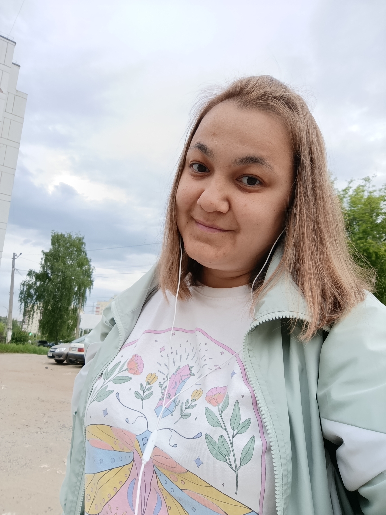A woman is taking a selfie outdoors. She has brown hair and is wearing a white t-shirt with a colorful graphic and a light green jacket. She also has white earphones plugged in.
Behind her are trees, a building, parked cars, and an overcast sky. The ground appears to be a dirt road or parking lot.
The image was taken in the city of Chaikovsky, Russia.