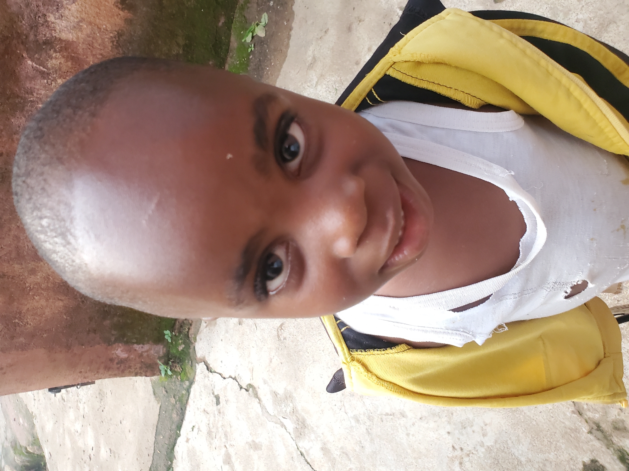 In Jos, Nigeria, a close-up, high-angle shot shows a young Black child from the chest up, looking directly at the camera. The child has a shaved head and is wearing a white tank top with visible holes and a yellow and black jacket draped over their shoulders. The background is a concrete surface with cracks and some greenery, adjacent to a reddish-brown textured wall.