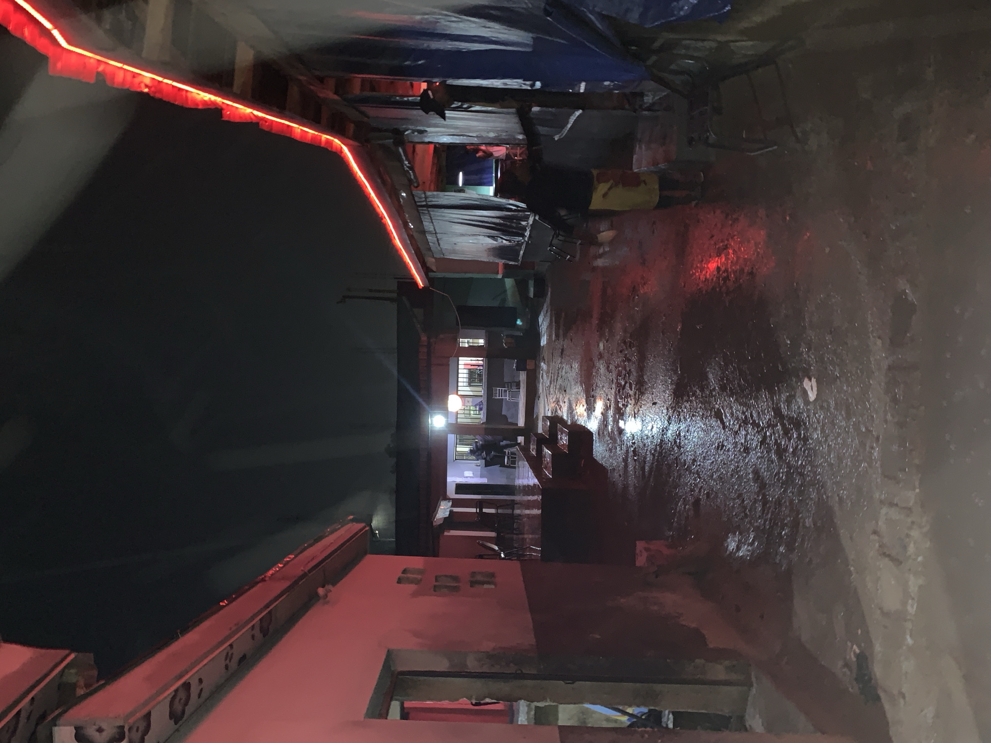 The image depicts a narrow alleyway at night. The ground appears wet, reflecting light from various sources. Above, a string of red lights stretches along the edge of a building or structure. Further down the alley, a building is visible with illuminated windows. The environment suggests an urban, possibly densely populated area, consistent with locations near New Stadium, Nigeria.