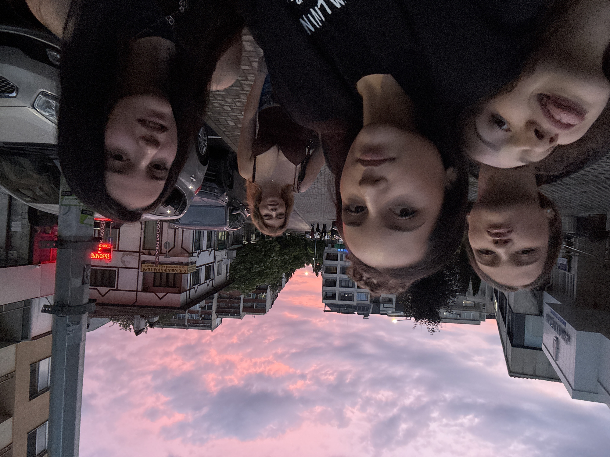 The image shows five young women posing for a selfie on a street in Unknown. The sky is a vibrant pink and purple, indicating either sunrise or sunset. The women are standing close together, with the camera held by one of them, creating a dynamic, close-up composition. The woman in the front left is sticking her tongue out. In the background, there are buildings lining the street, some with shops and signs. Cars are parked along the side of the road. The overall atmosphere is casual and friendly.