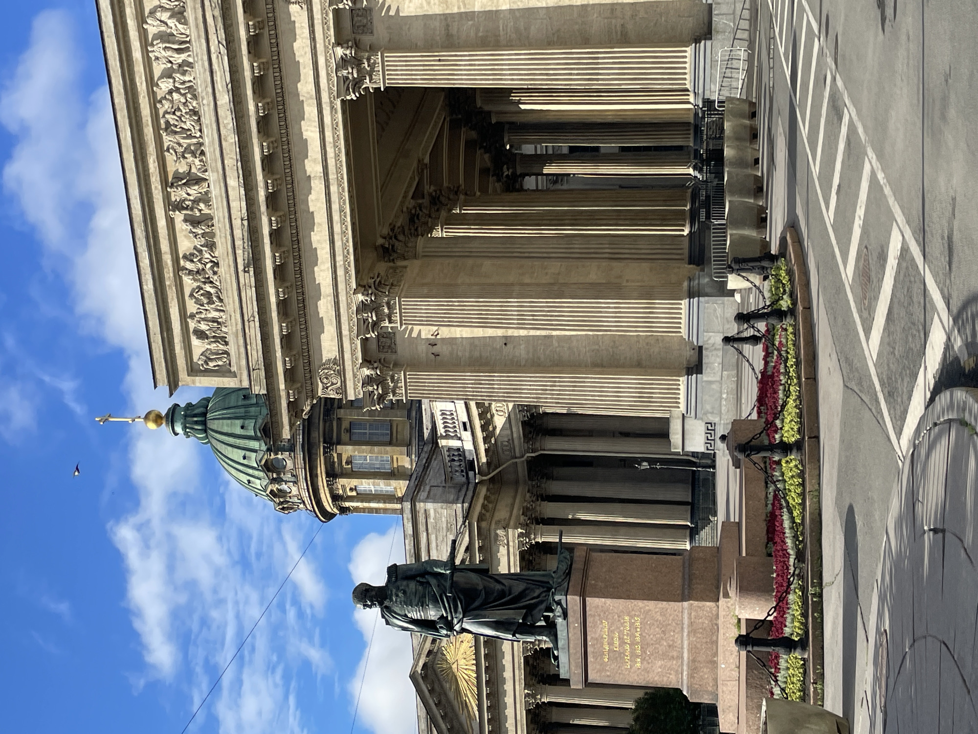 The image shows the Kazan Cathedral in Saint Petersburg, Russia, on a clear day with scattered clouds. In the foreground, a statue of a man in military attire stands on a pedestal. The statue is dark bronze and the pedestal is a light reddish-brown with inscriptions in gold lettering. A circular flower bed with red and yellow flowers is visible in front of the statue, bordered by a black metal chain fence. The Kazan Cathedral's grand colonnade, made of large, fluted columns, extends to the right of the statue. The cathedral's dome, topped with a golden cross, is visible in the background. The sky is blue with white clouds. The ground in the foreground appears to be paved with concrete and asphalt, with white lines indicating a pedestrian crossing.